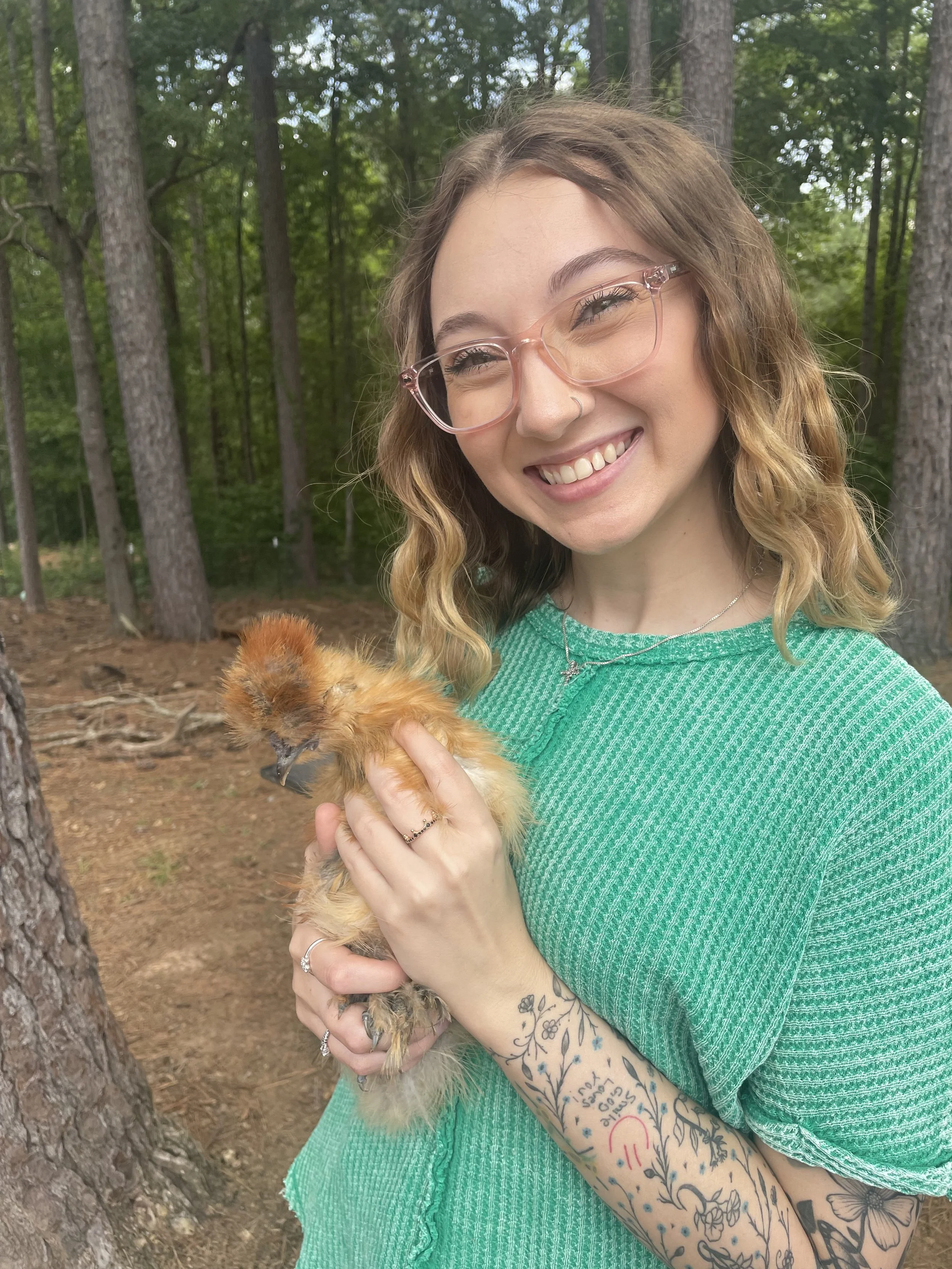 A young woman with glasses, blond wavy hair, and tattoos on her arm holds a fluffy chicken outdoors in a wooded area.