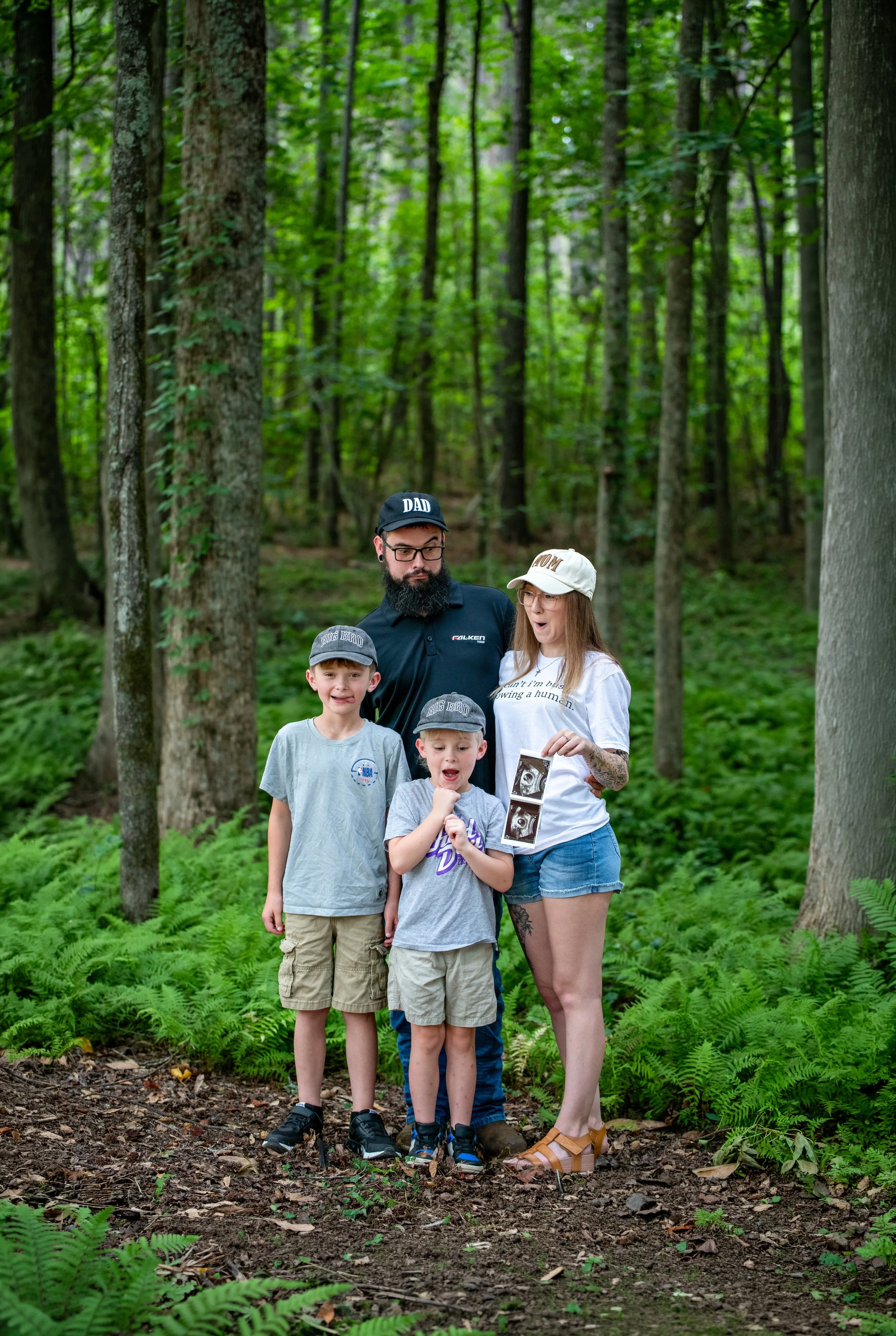 Family of four standing among trees and ferns in a lush forest, with the mother holding ultrasound photos.
