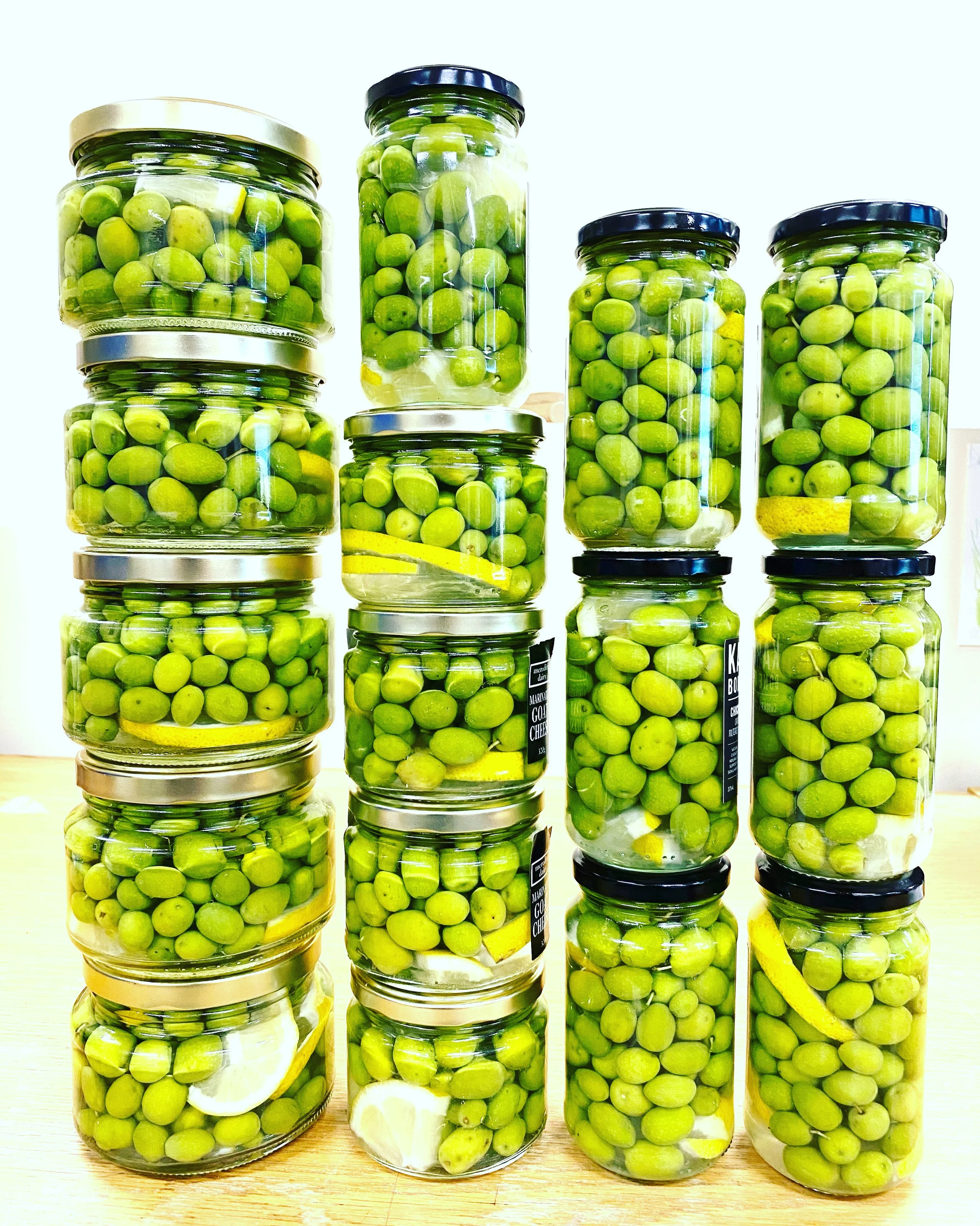 Multiple jars filled with green olives, some with lemon slices and herbs, arranged on a wooden surface.