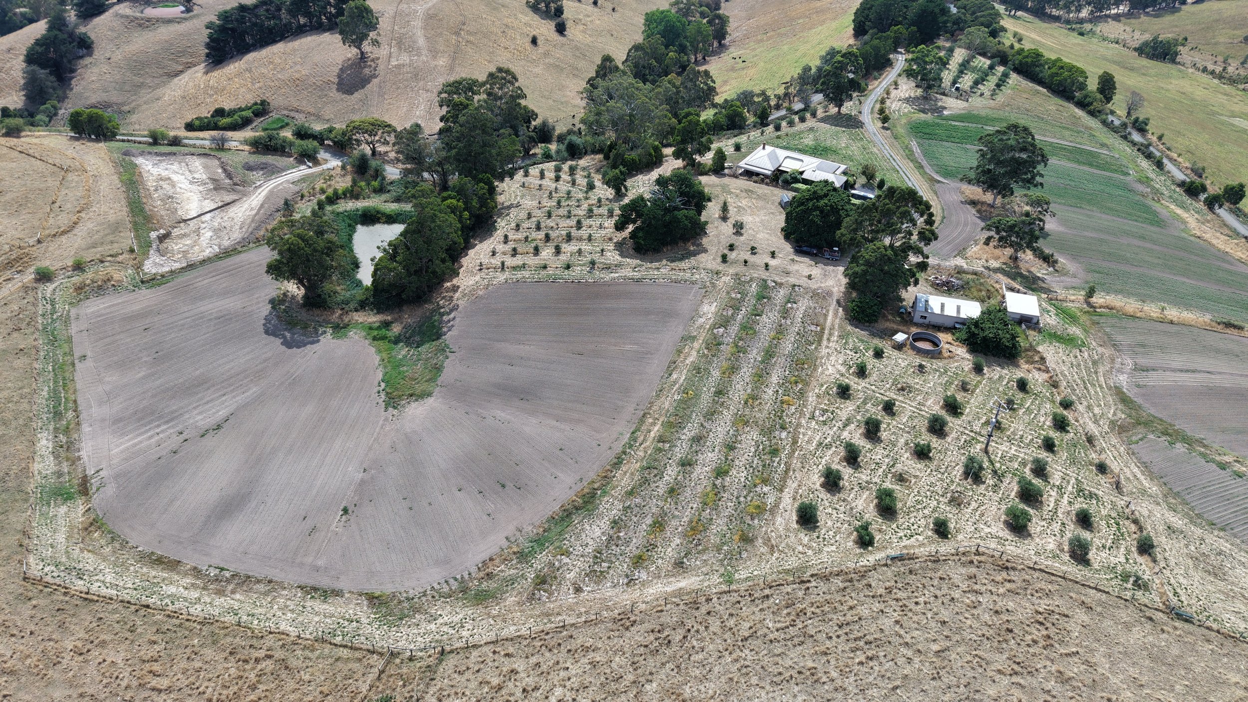 Aerial view of a rural landscape with farmland, trees, and a few houses, including a pond and dirt roads.