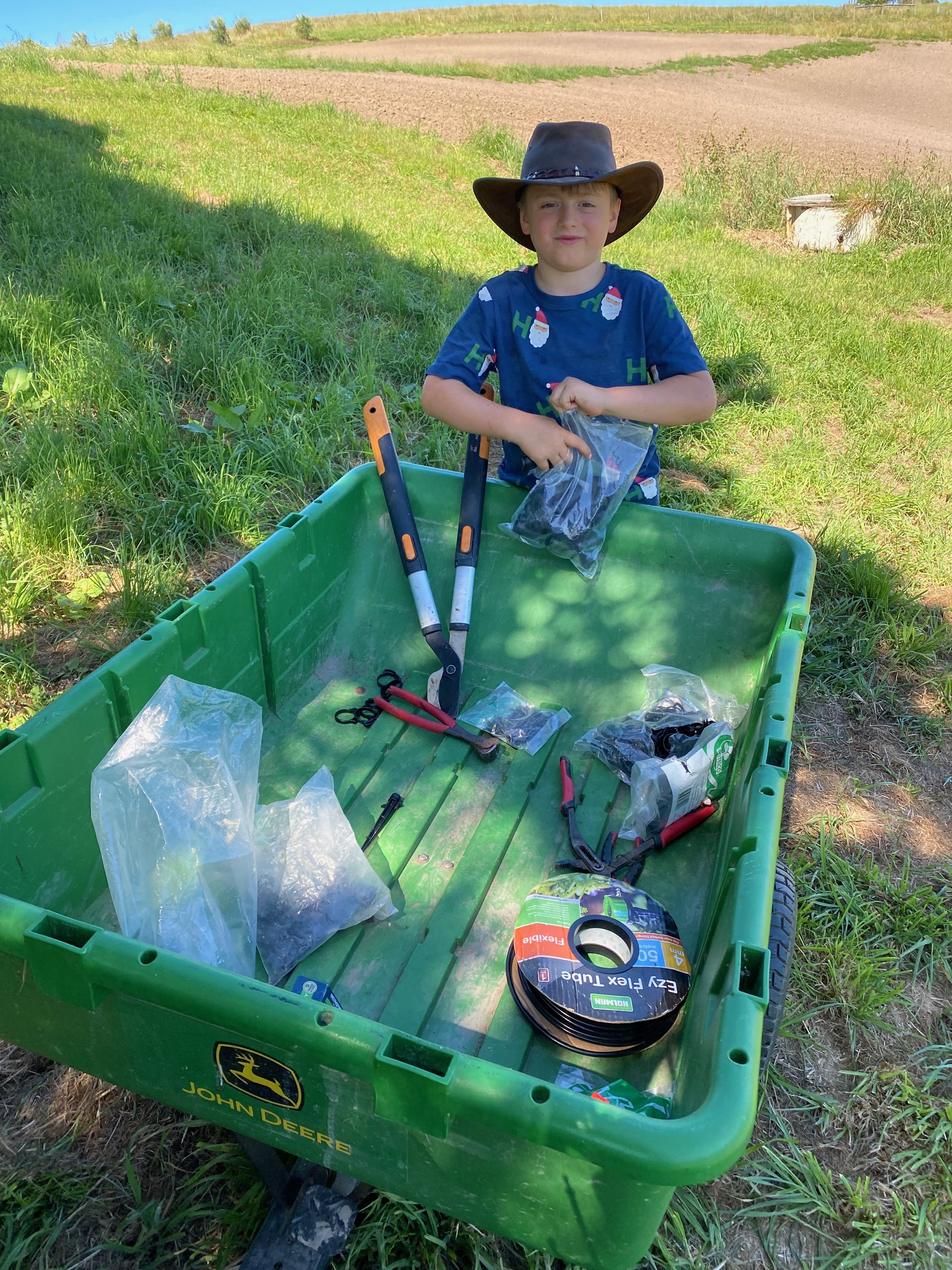 A young boy wearing a large black cowboy hat and a blue shirt with Santa Claus and holiday patterns, sitting next to a green John Deere utility cart filled with gardening or yard work tools, outdoors on a grassy and dirt area with farmland in the bac
