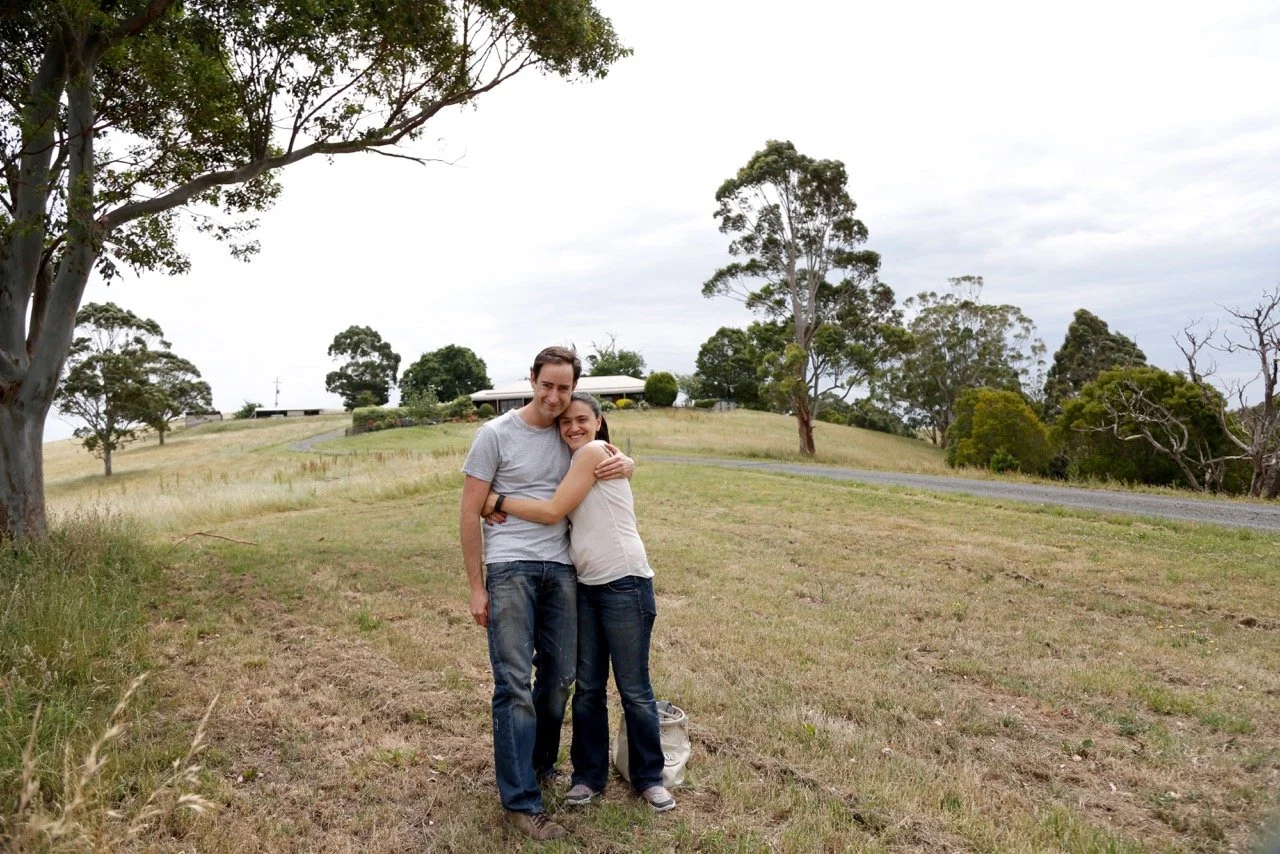 A young man and woman hugging outdoors in a grassy field with trees and a house in the background, overcast sky.