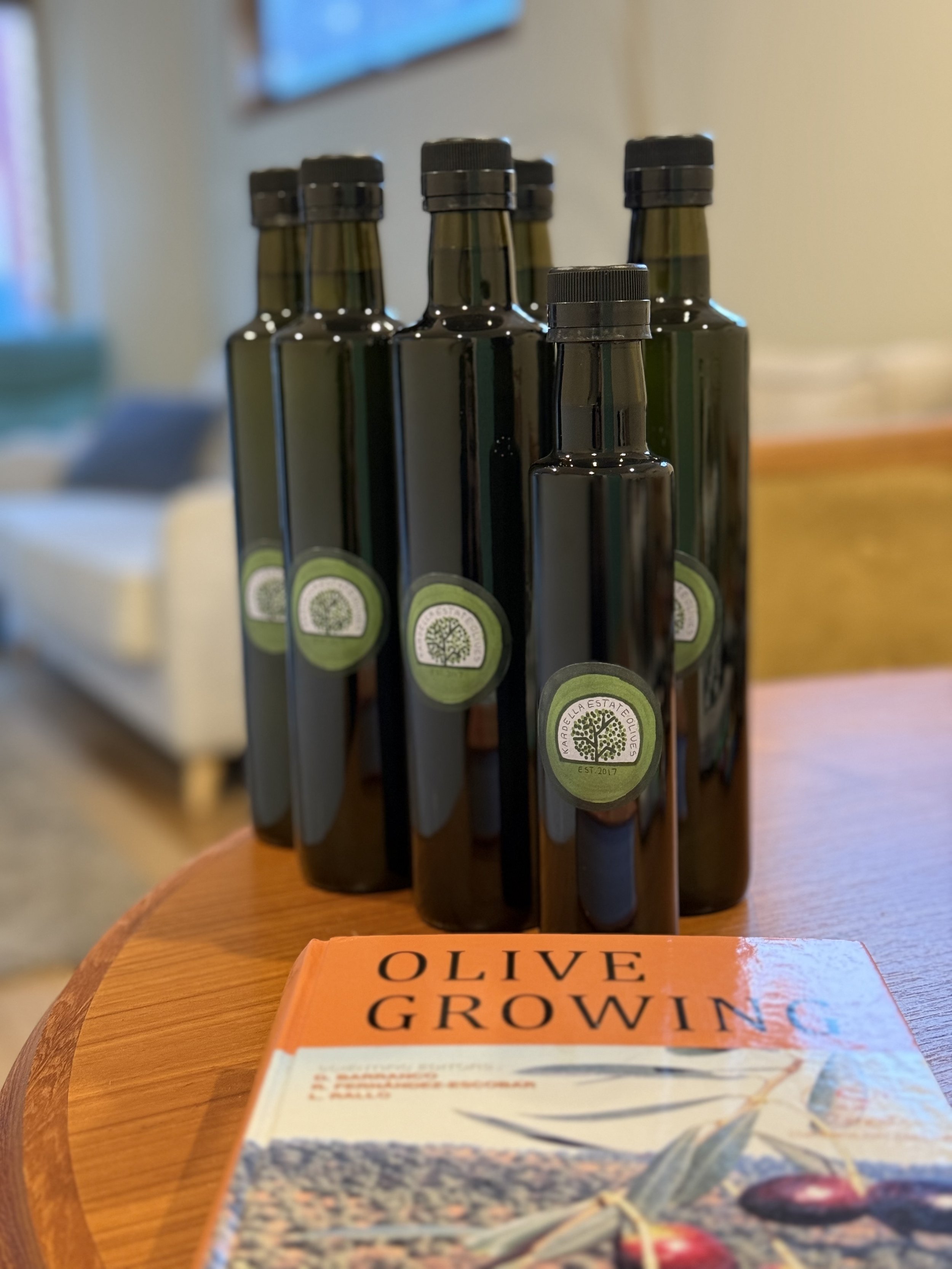 Five dark green glass bottles with black caps and a label featuring a tree logo are arranged upright on a wooden surface in a staggered formation, with a book titled "Olive Grown" resting in the foreground. The background is softly blurred.
