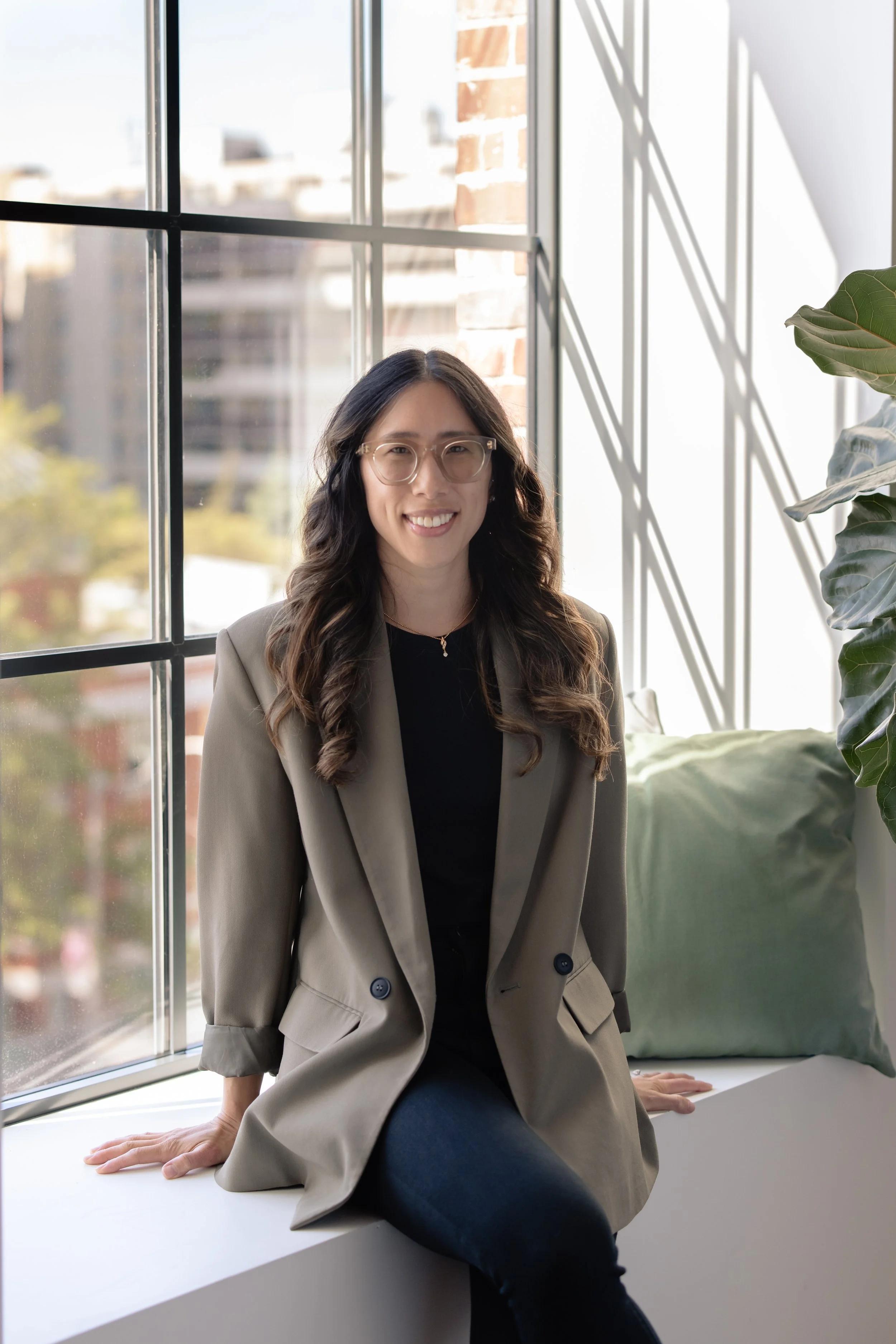Image of woman sitting in a window sill with a green pillow and plant to her left.