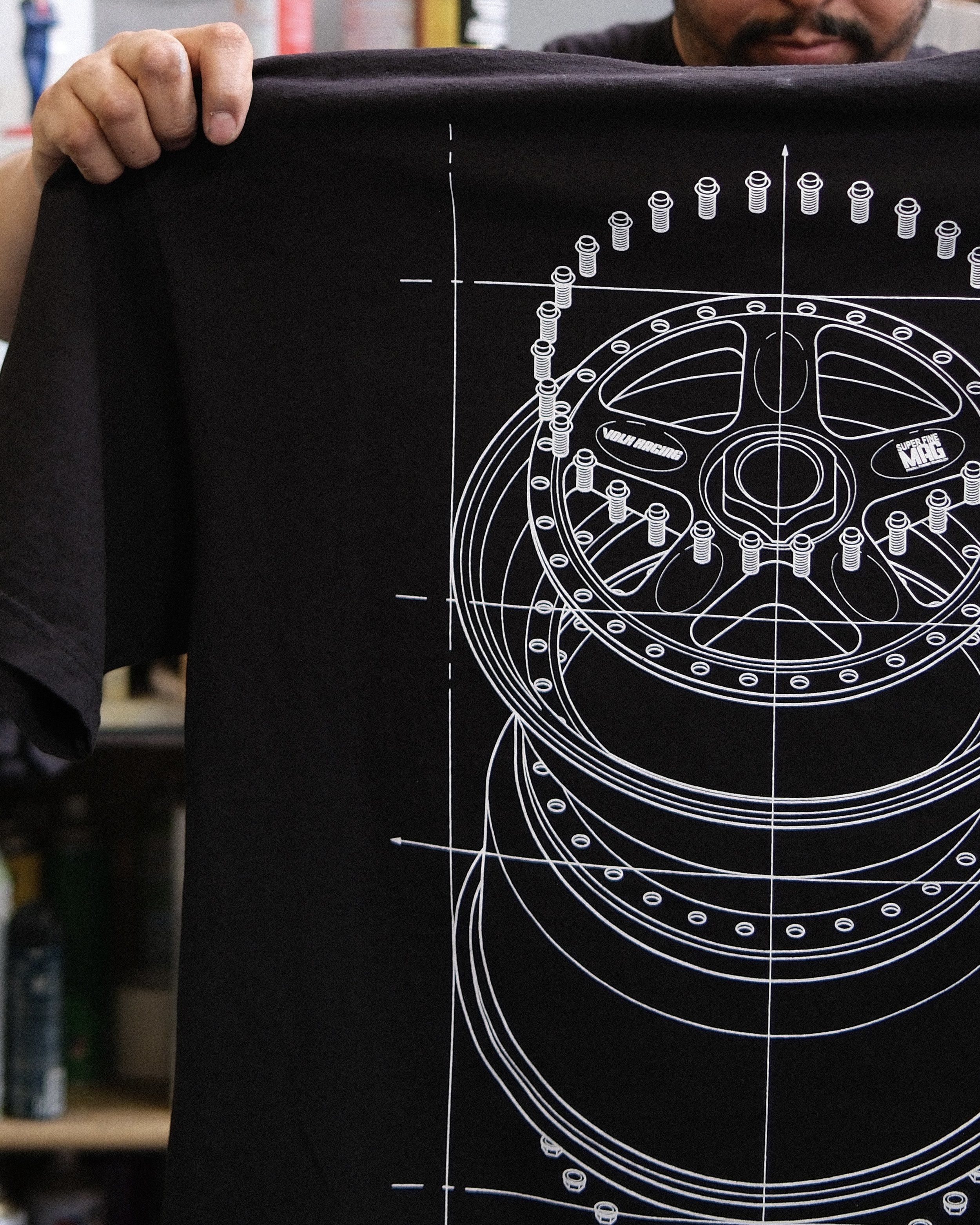 A man holding up a black T-shirt with a white technical drawing of a wheel and bolt pattern.
