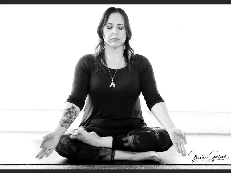 A woman practicing meditation in a seated cross-legged pose, eyes closed, in black and white, with tattoos on her arms and a pendant necklace, sitting against a plain background.