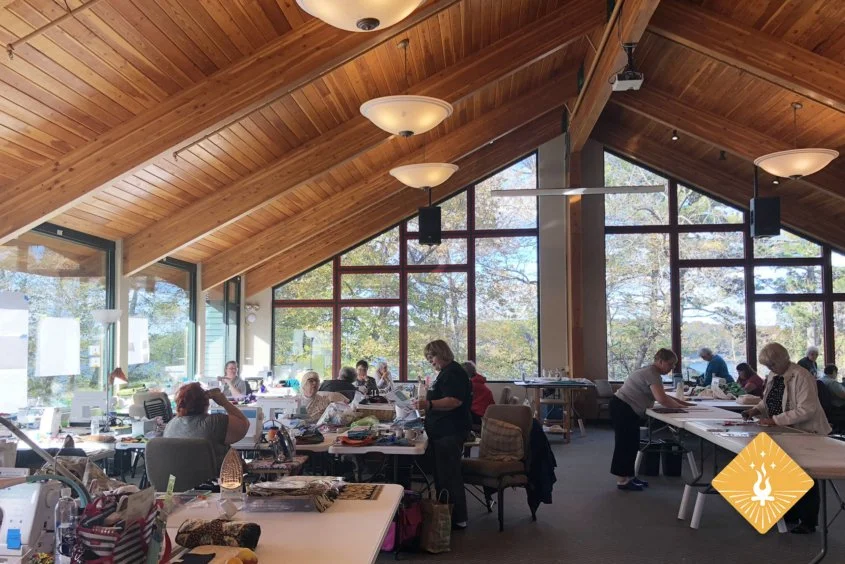 People gathered in a spacious room with large windows, working on craft projects at tables under a wooden, vaulted ceiling with ceiling lights.