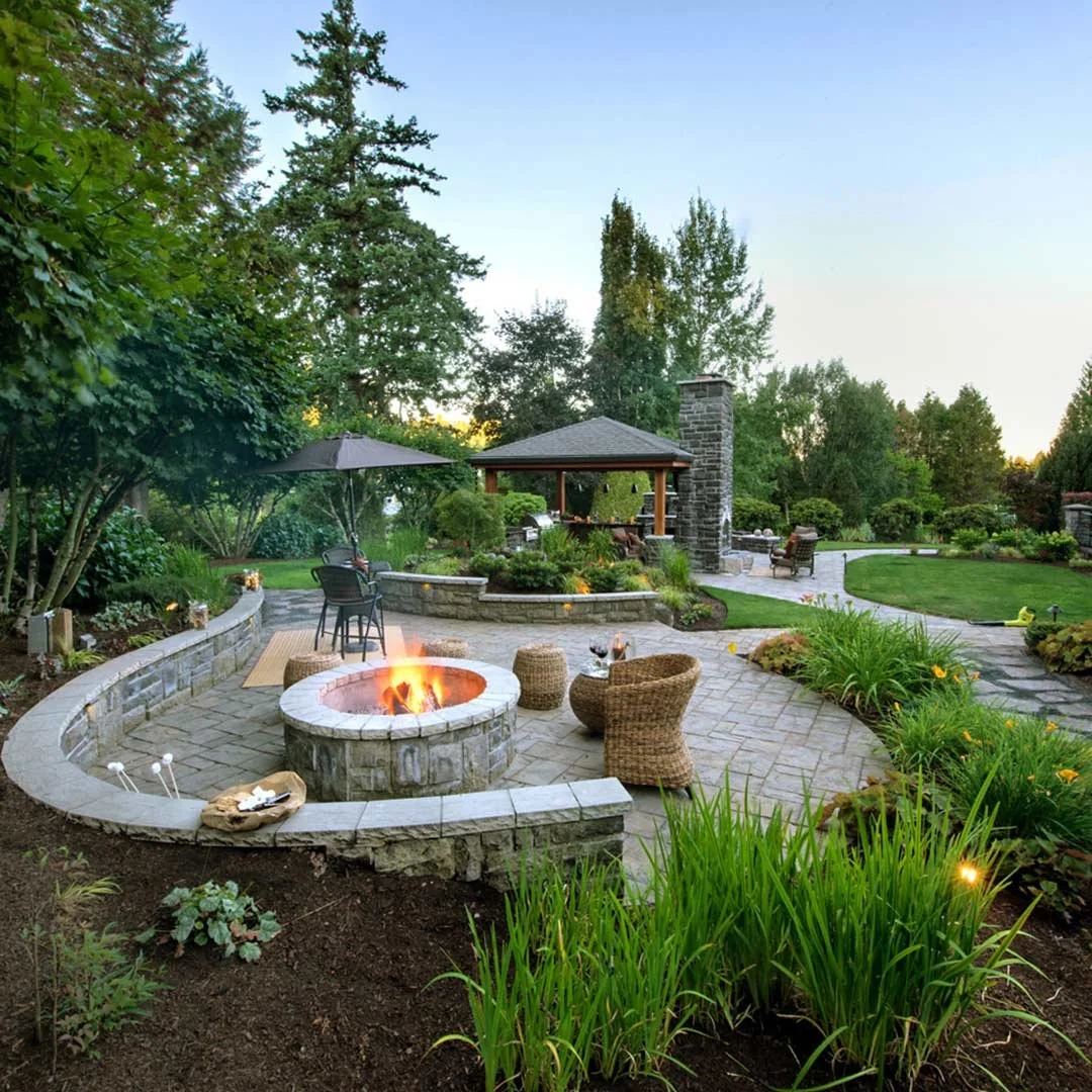 A landscaped backyard featuring a stone fire pit with chairs, a gazebo, a patio, lush greenery, trees, and outdoor furniture, all illuminated by soft lighting during dusk.