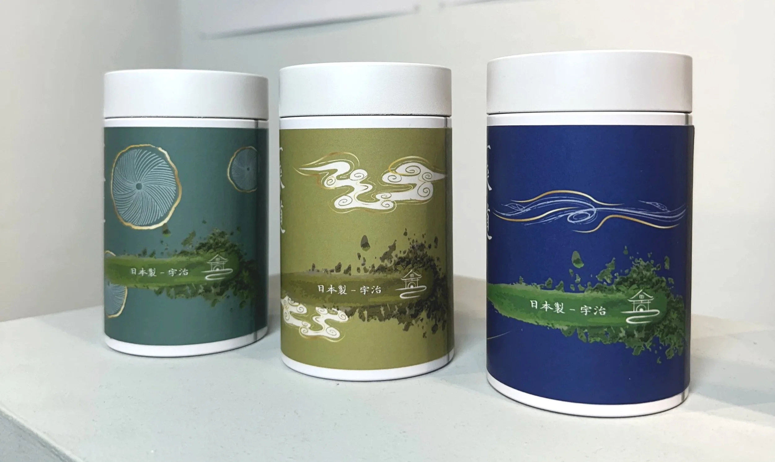 Three cylindrical containers with colorful labels featuring Japanese characters and illustrations of clouds, trees, and landscapes, placed on a white surface against a plain background.