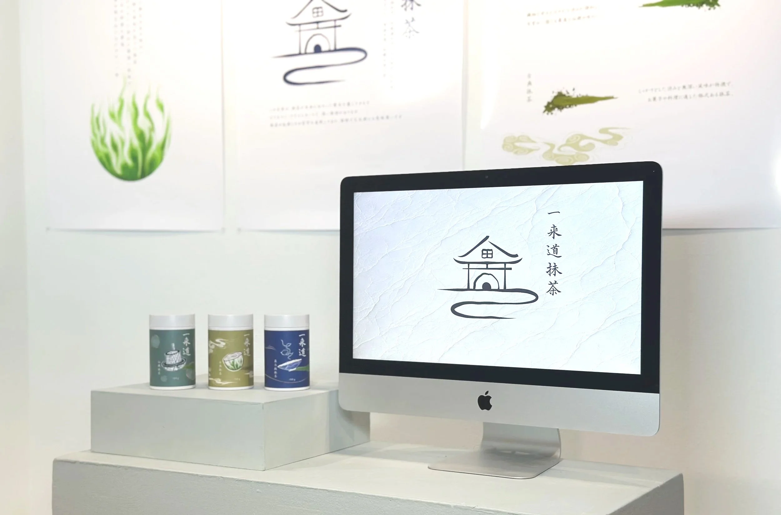 An Apple iMac computer on a white surface displaying a logo with a simple line drawing of a traditional Asian building and Chinese characters. Behind the computer, there are three cans with different colors and designs, placed on a slight elevation. In the background, there are three posters on the wall, each featuring minimalist artwork and Chinese text.