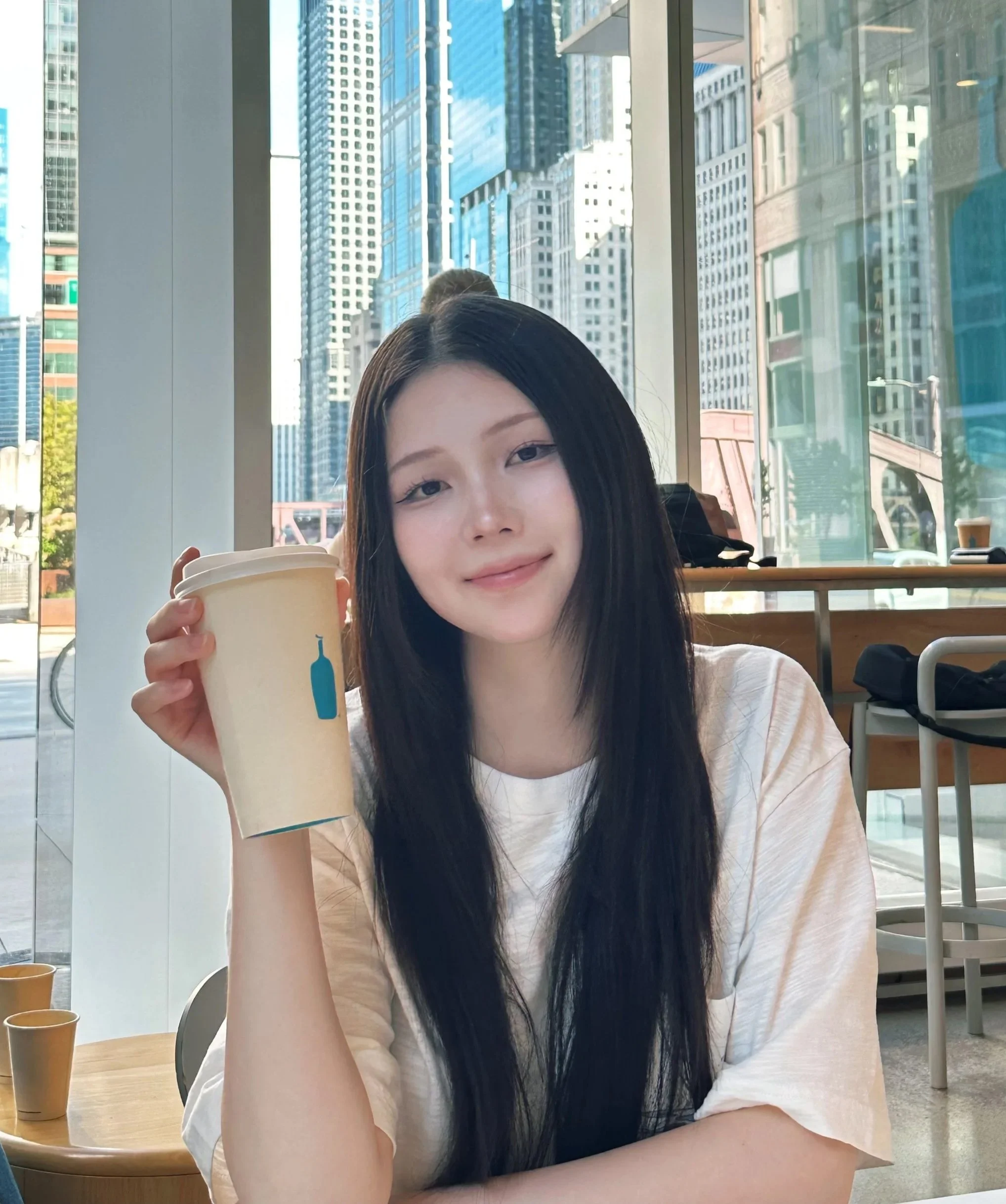 A young woman with long black hair holding a disposable coffee cup inside a cafe with large windows showing skyscrapers outside.