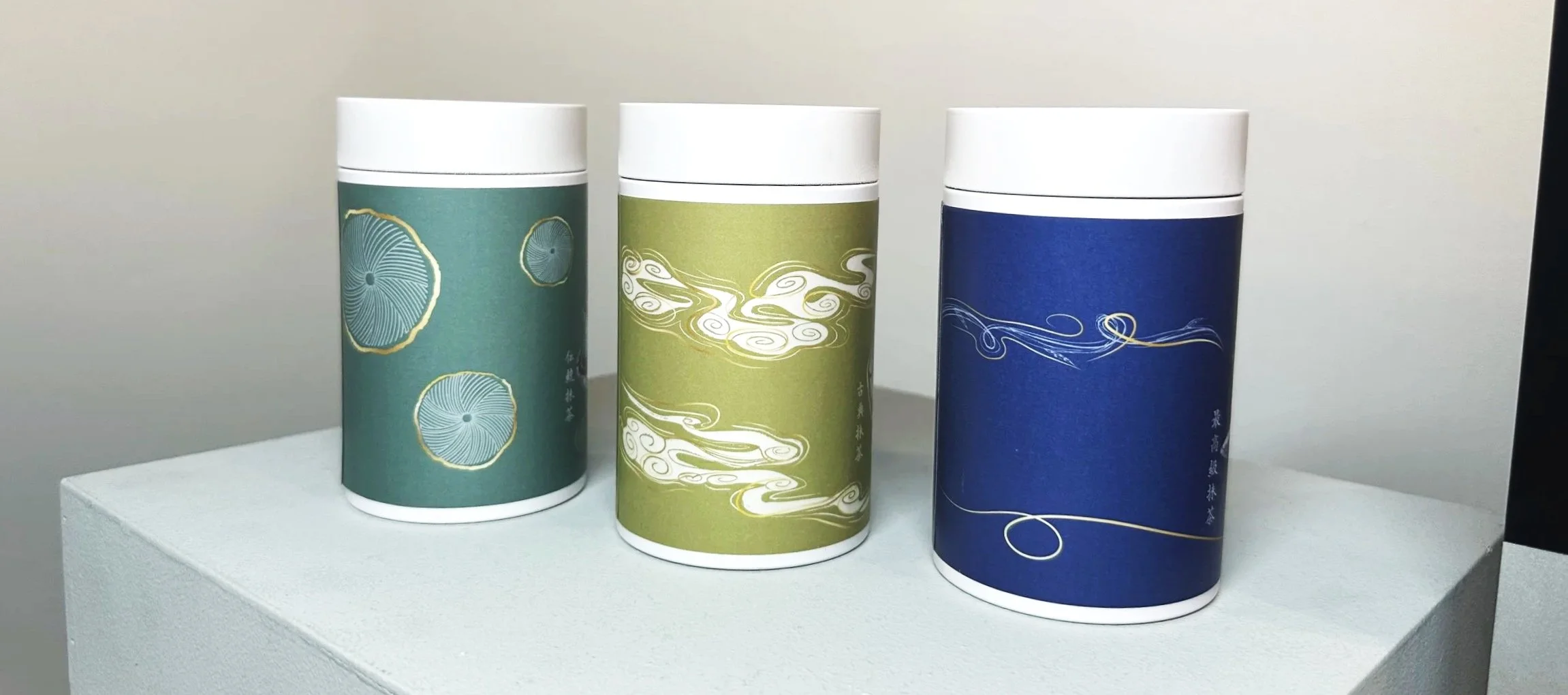 Three cylindrical containers with simple, elegant Japanese-style drawings in blue, white, and green colors, placed on a white pedestal against a plain background.