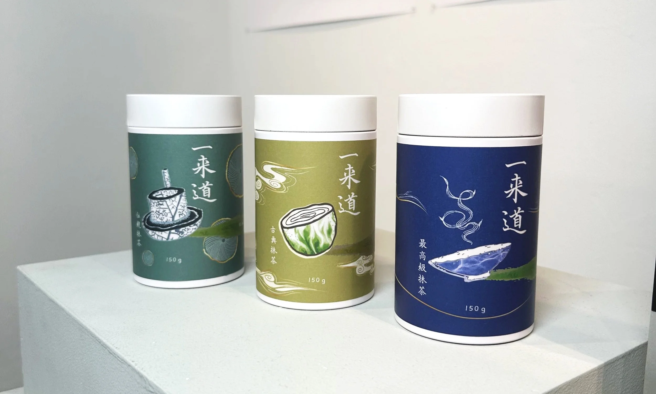 Three cylindrical containers with Japanese characters, each featuring a different color and illustration of matcha-related items, placed on a white surface against a white background.