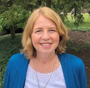 A woman with blonde hair wearing a blue cardigan and white top, smiling outdoors with trees in the background.
