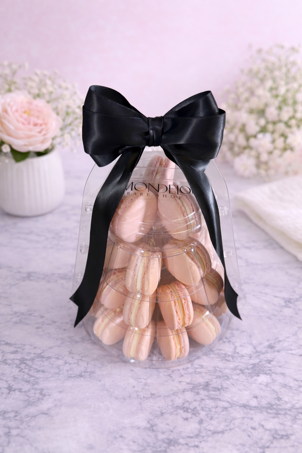 A clear plastic container filled with pink macarons decorated with a large black satin bow on top, placed on a marble surface with flowers in the background.