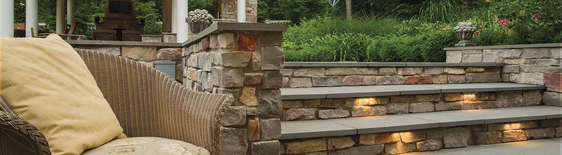 Outdoor stone staircase with built-in lighting next to a garden and wicker furniture with cushions.