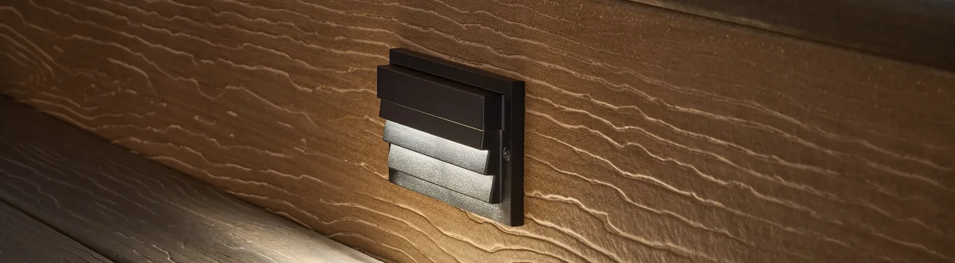 Close-up of a modern black outdoor wall light fixture mounted on a textured wooden wall, casting a soft glow downward.