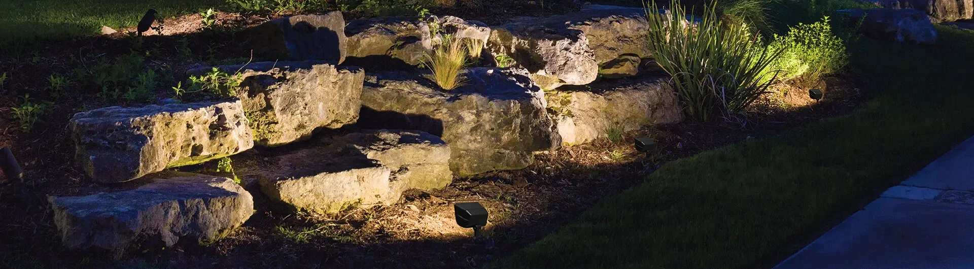 Rock garden with landscape lighting at night, featuring various rocks, plants, and grass.