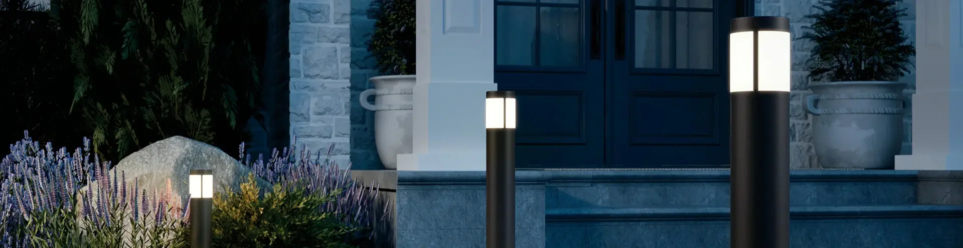 Nighttime exterior view of a house with a blue door, stone steps, large white flower pots, and garden plants. Three outdoor pathway lights illuminate the scene.