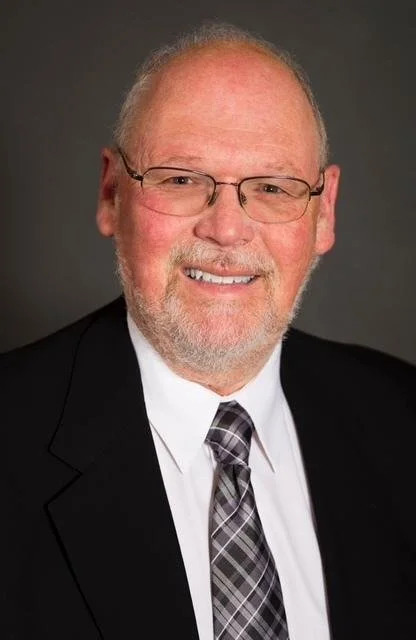 A middle-aged man with light skin, glasses, a beard, and a smile, wearing a black suit, white shirt, and a patterned tie, against a dark background.