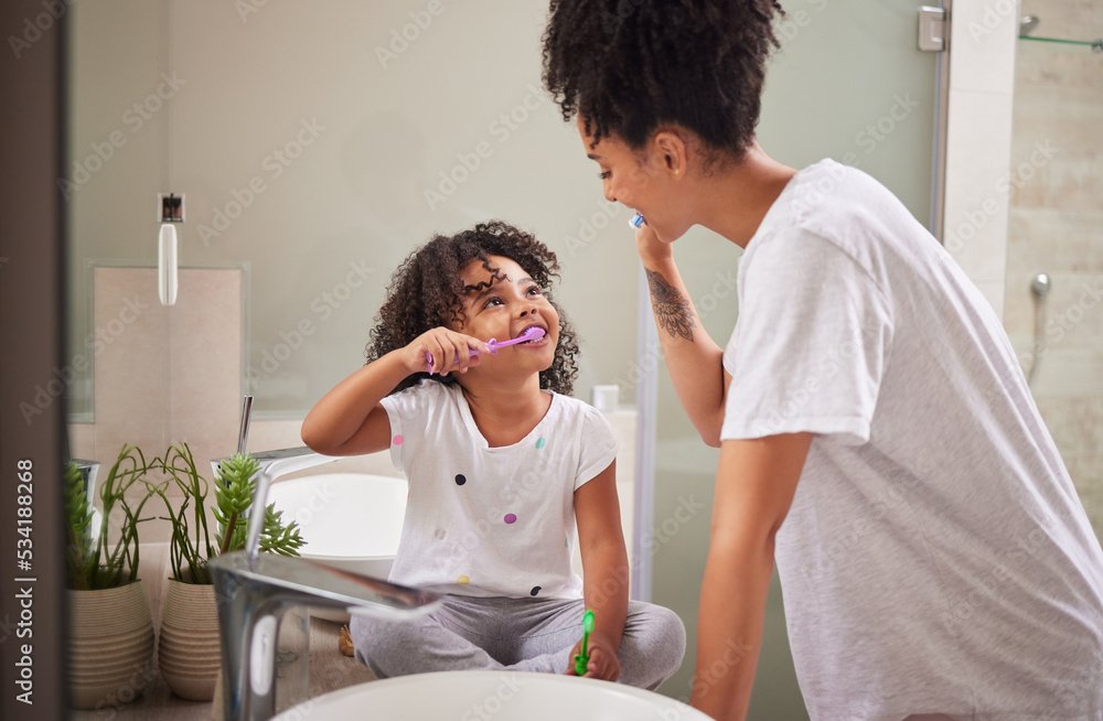 A young girl brushing her teeth in front of a mirror with her mother leaning over to assist or talk to her at the bathroom sink.