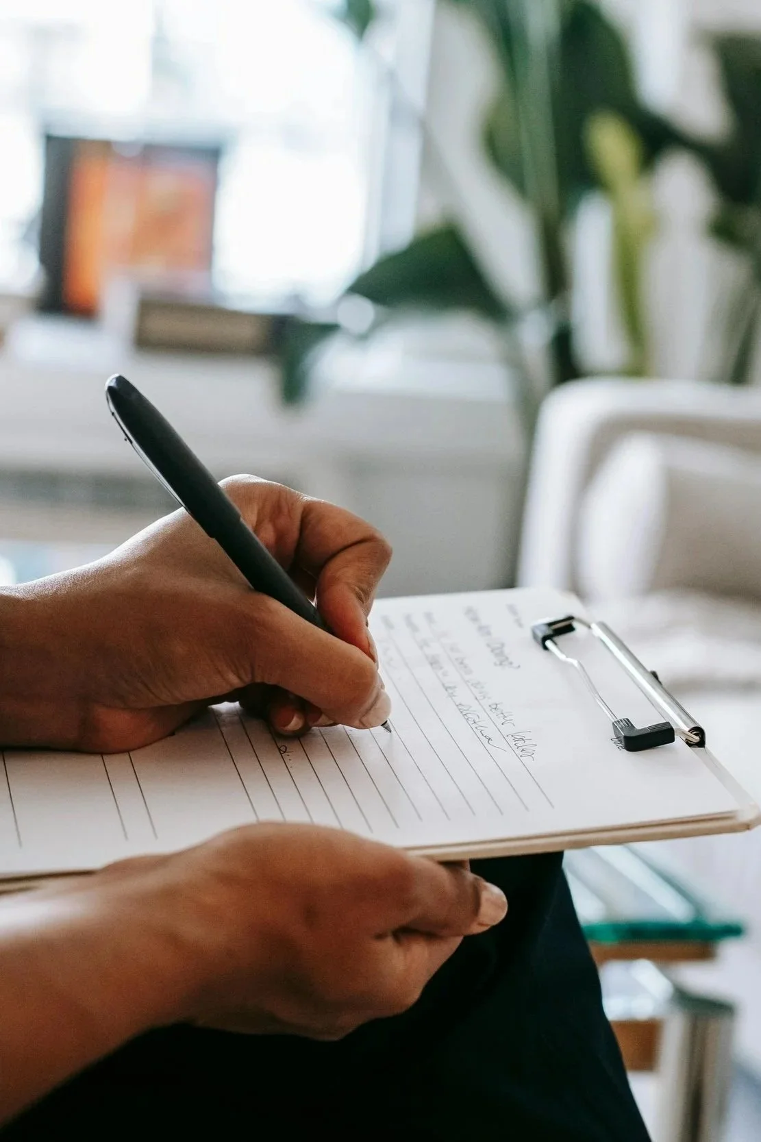 Person writing with a pen on a clipboard in a well-lit room with plants and a window in the background.