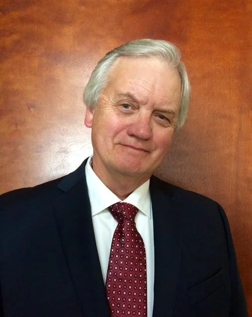 A middle-aged man with gray hair in a suit and red tie, standing in front of a wooden-paneled wall.