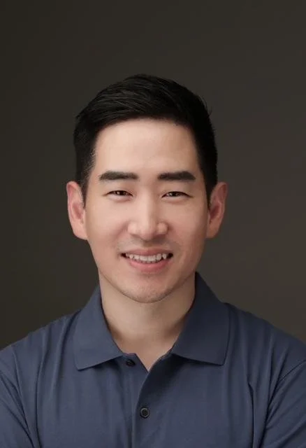 A young man with short black hair smiling, wearing a navy blue collared shirt, against a dark gray background.