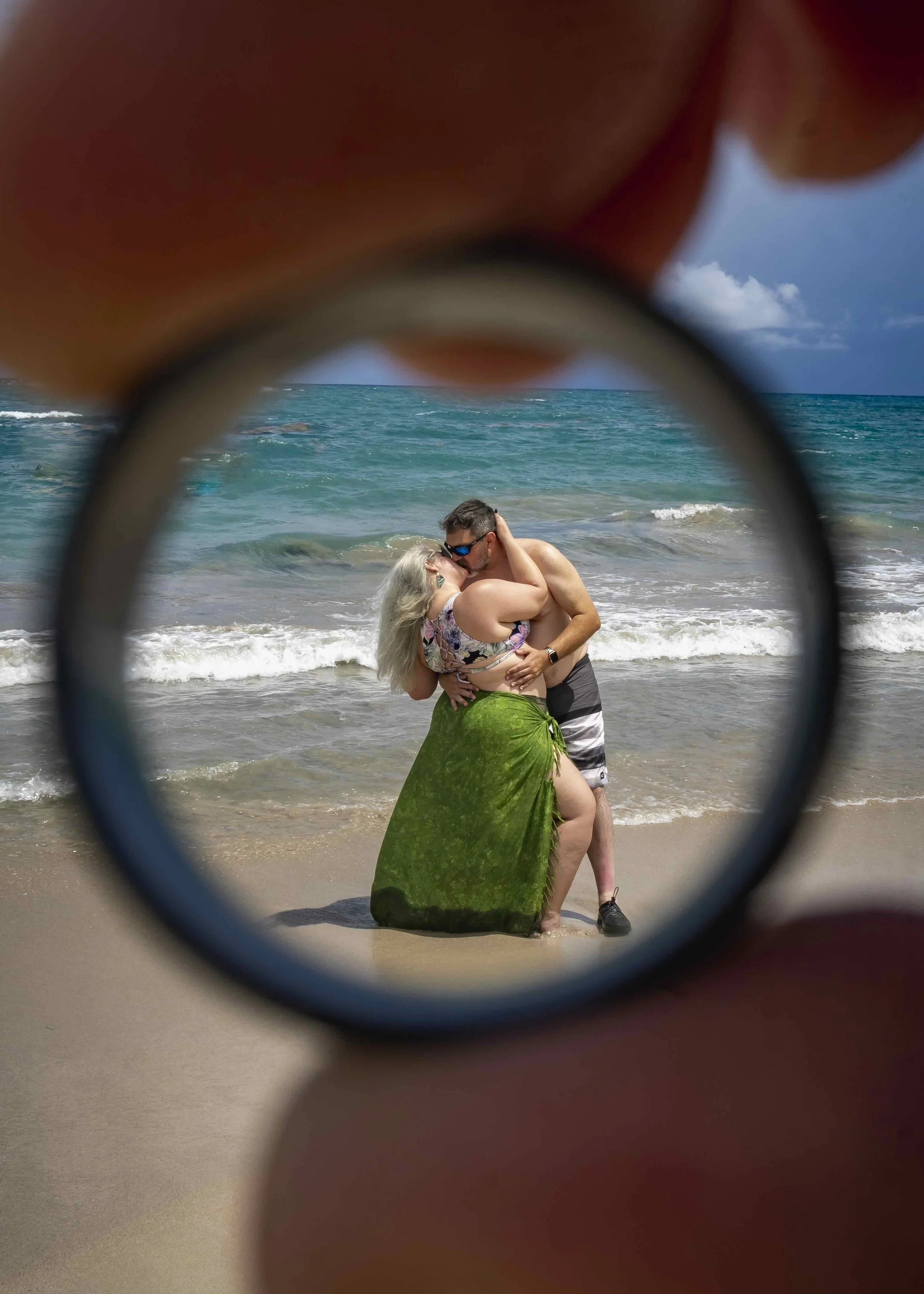 A couple kissing and embracing on a beach, viewed through a wedding ring with the ocean and sky in the background.