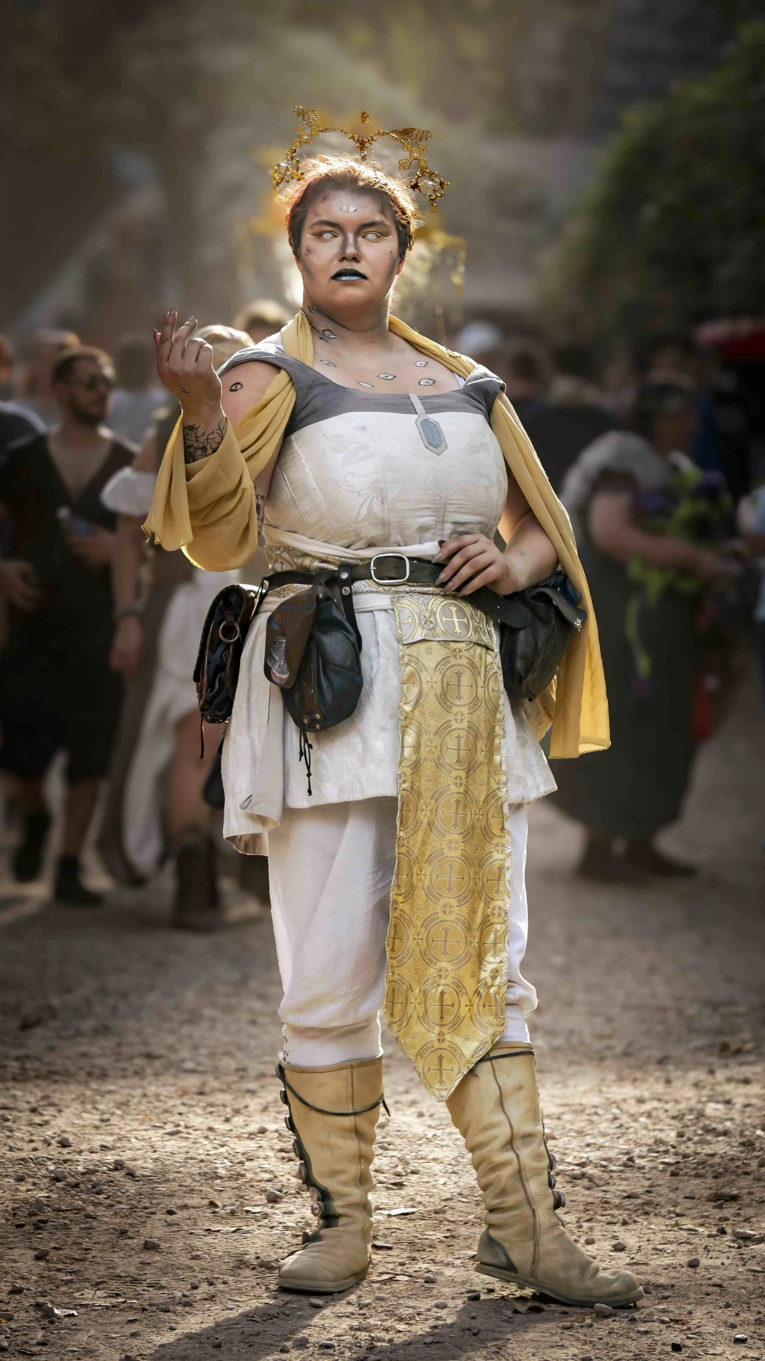 Person wearing a fantasy or cosplay costume with a crown-like headpiece, face makeup, tattoos, and clothing resembling historical or fantasy armor, standing outdoors on a dirt ground with group of people in the background.