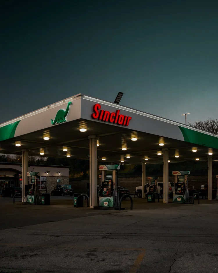 A Sinclair gas station at dusk with multiple fuel pumps under a canopy featuring green and white colors, a green dinosaur logo, and illuminated lights.