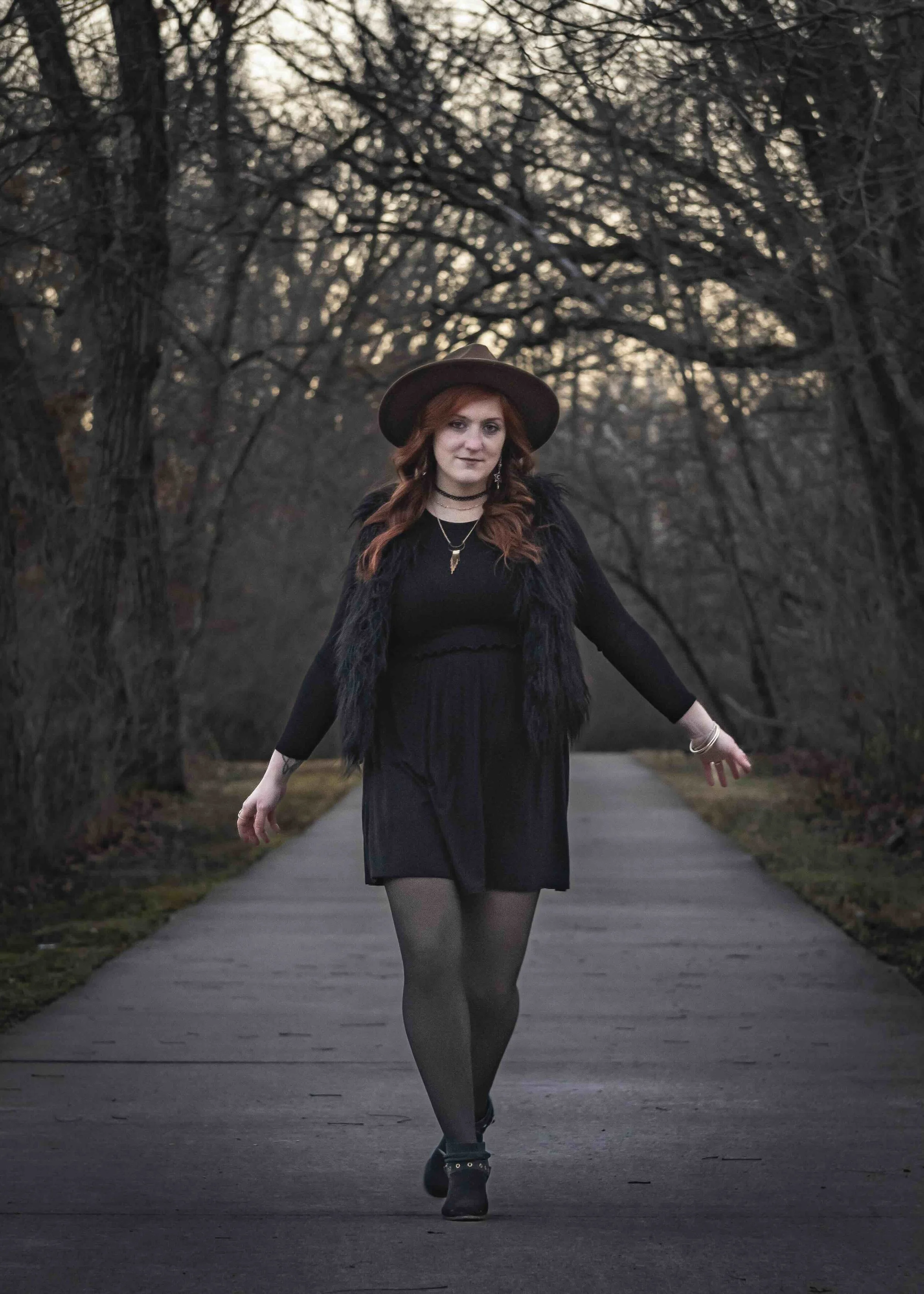 A woman with red hair walking on a paved path in a wooded area during dusk, wearing a black dress, a hat, and black tights.