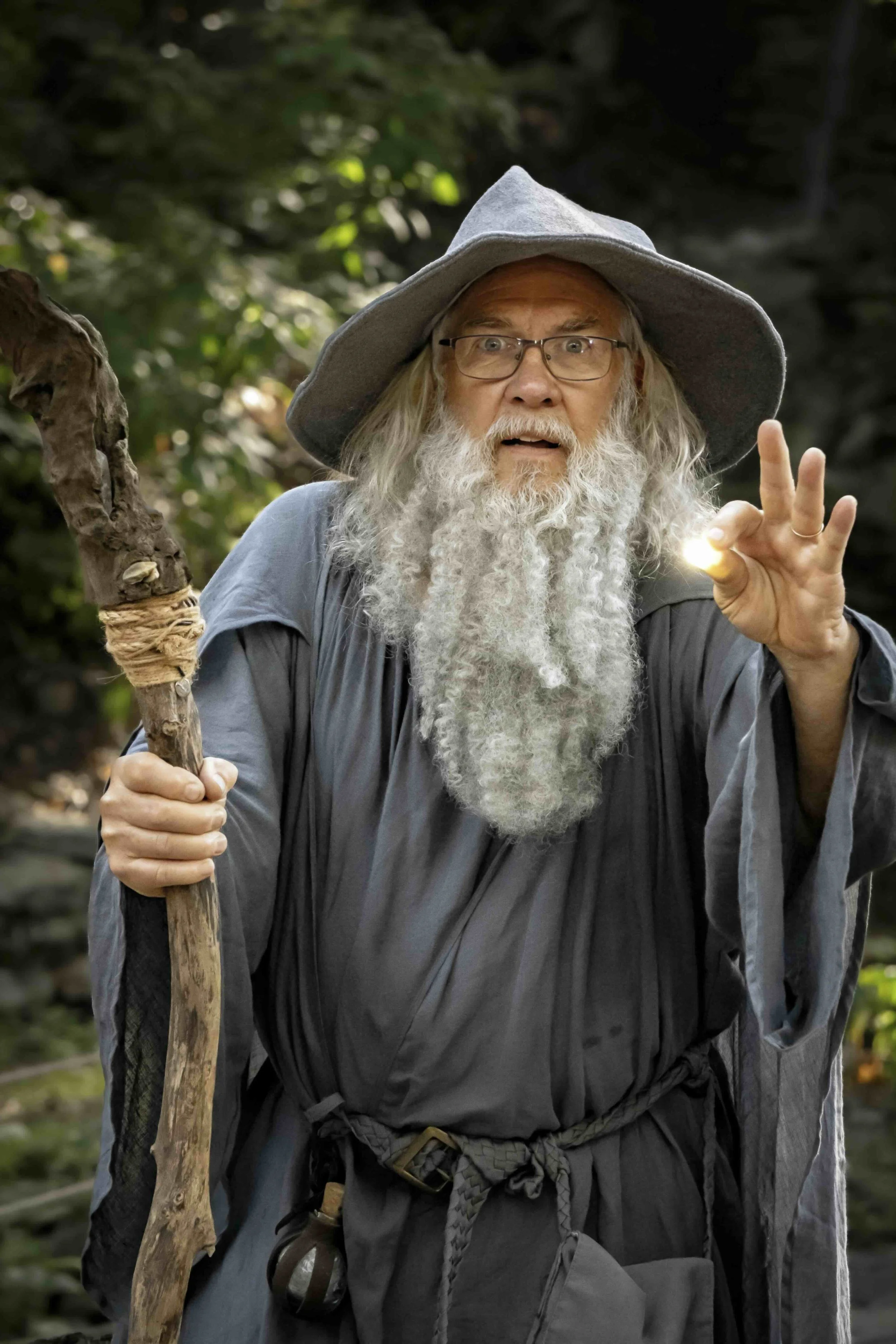 An elderly man dressed as a wizard with a long white beard and glasses, holding a wooden staff, in a forest, casting a spell with his hand.