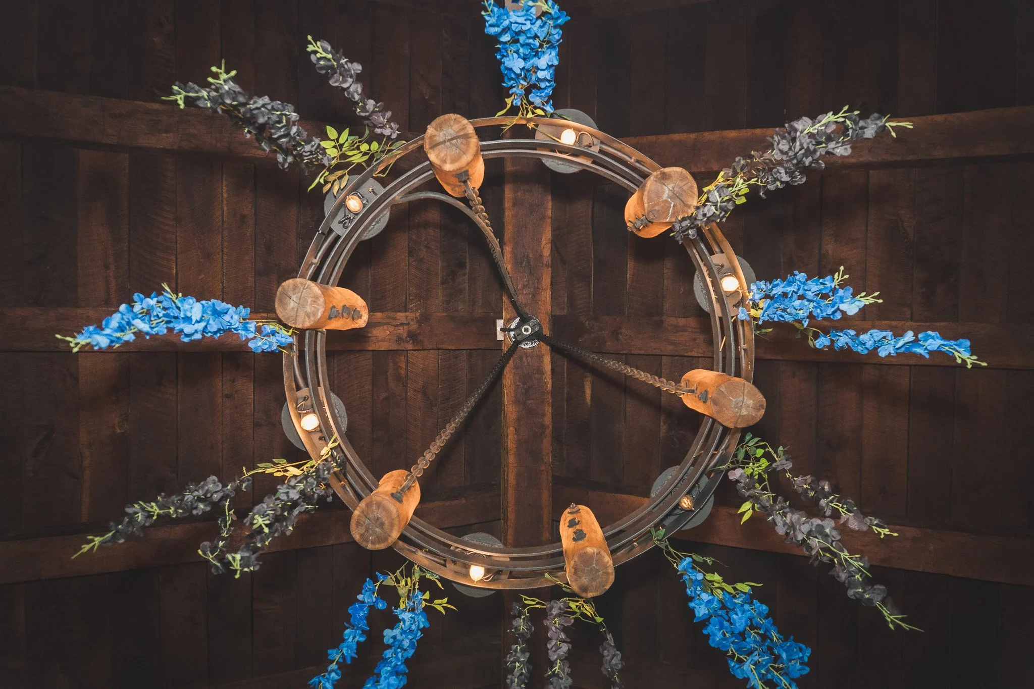 A round chandelier with metal rings decorated with blue and purple artificial flowers and small lights, hanging from a dark wooden ceiling.