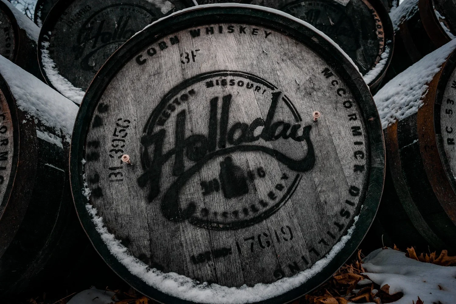 Wooden bourbon whiskey barrel with 'Holladay' handwritten on the side, partially covered in snow, stacked outdoors on the ground.