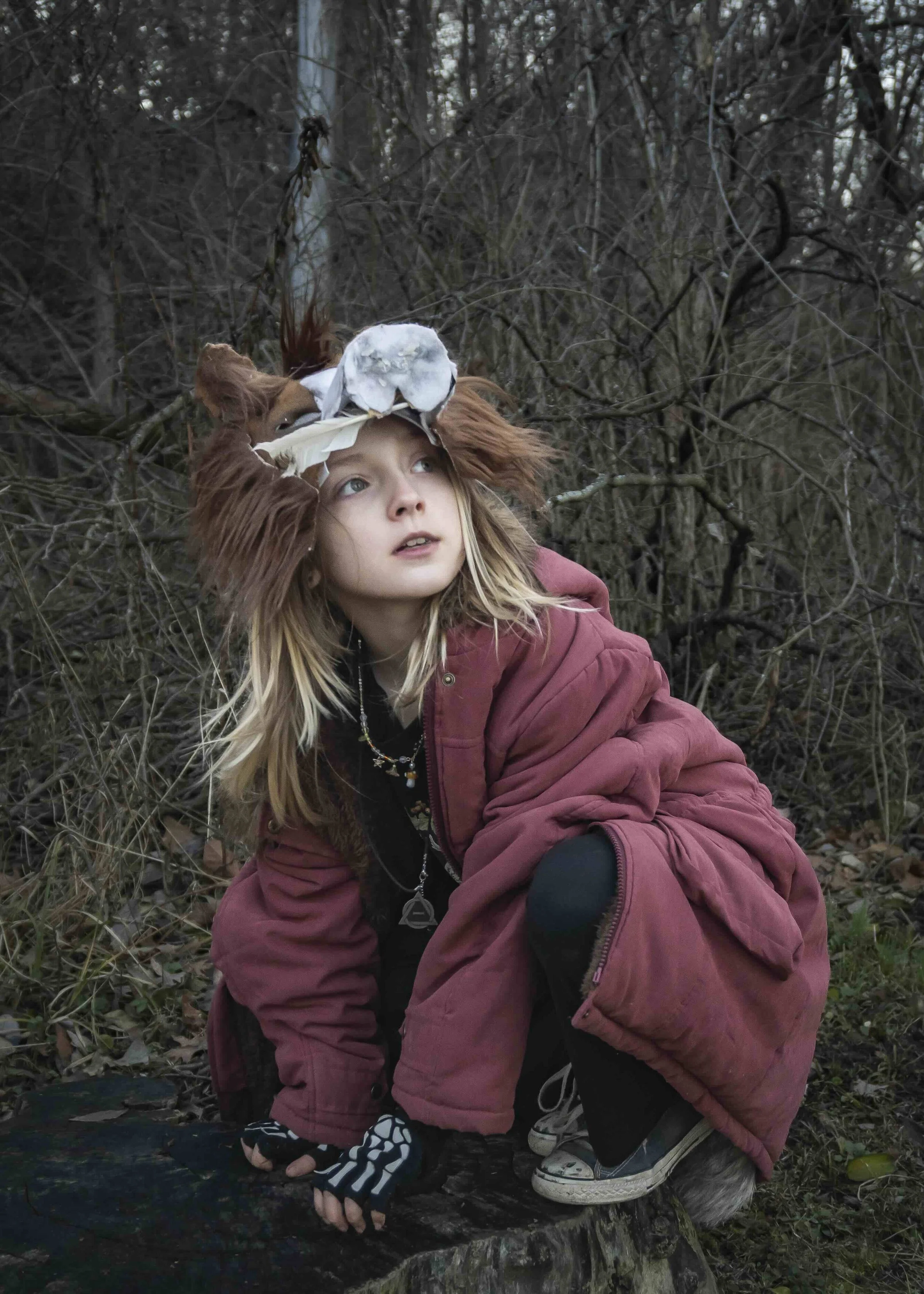 Young girl wearing a bird skull headdress and a red jacket crouching on a tree stump in a wooded area with tangled branches in the background.