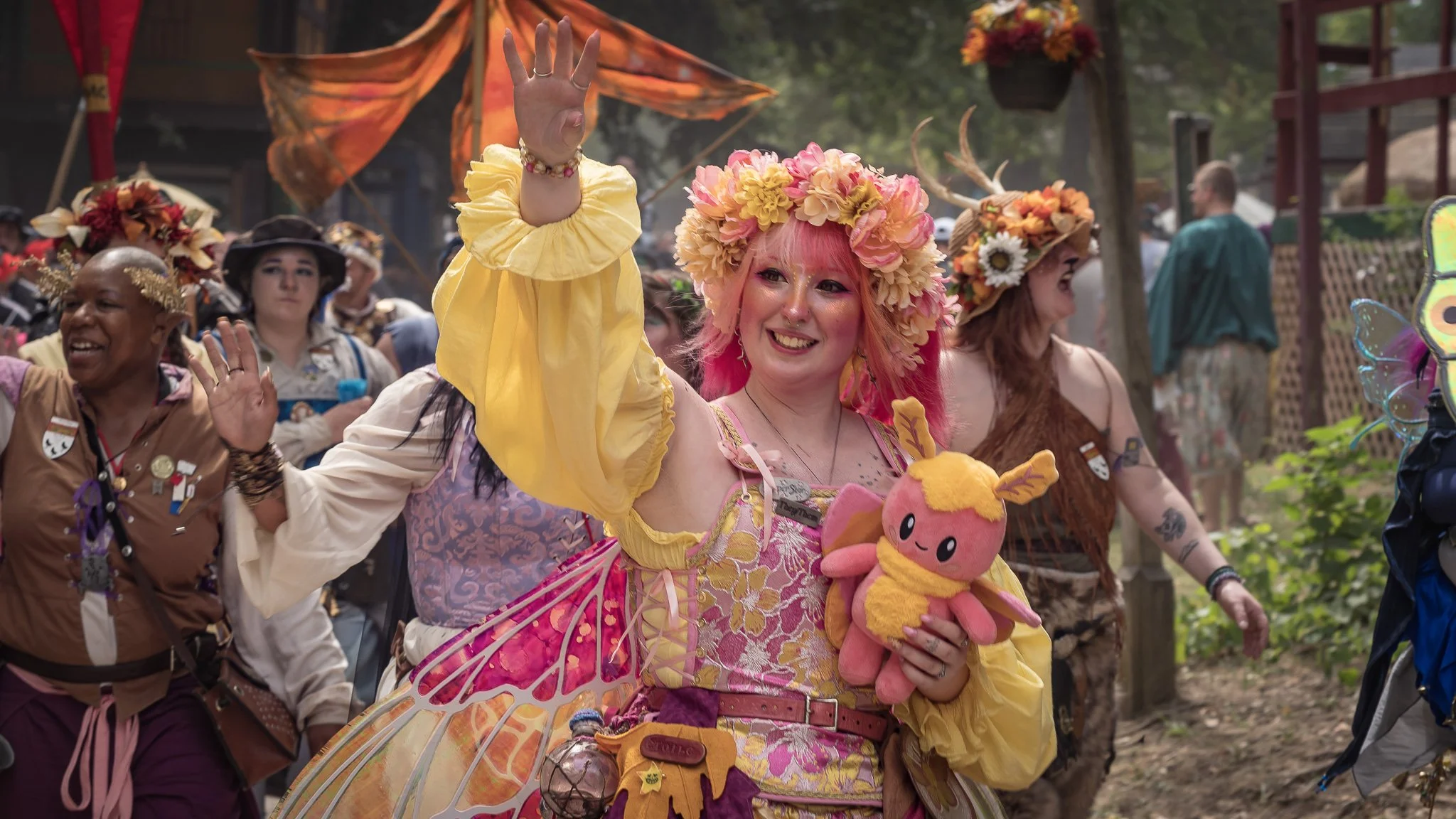Person dressed as a fairy princess with pink hair, adorned with a flower crown, holding a plush toy, and surrounded by others in colorful costumes at a festival or parade.