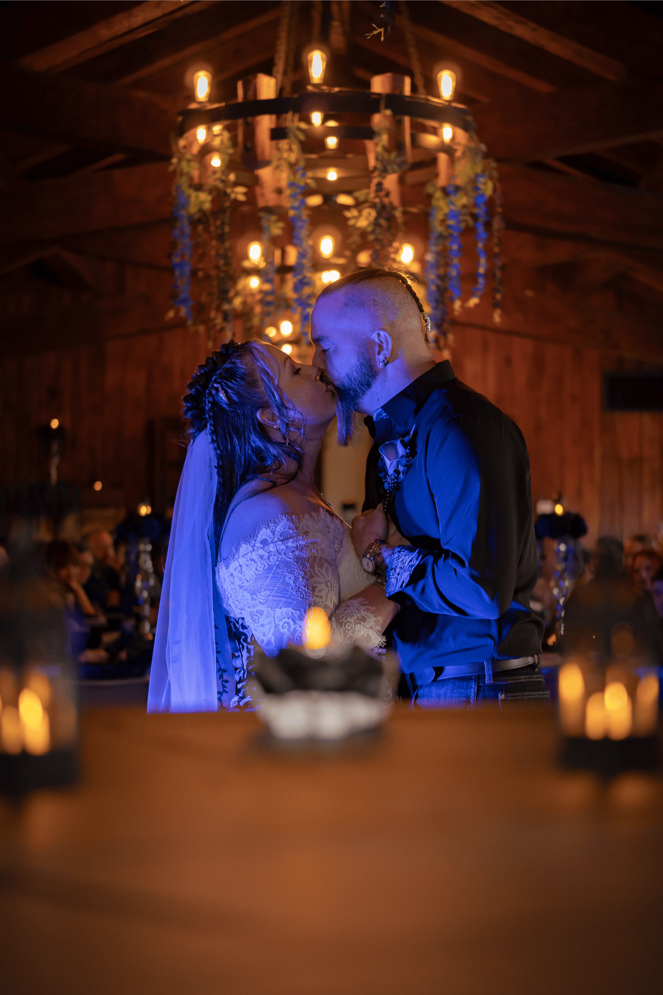 A bride and groom share a kiss during their wedding ceremony in a warmly lit wooden venue, with guests seated in the background and candles on the table in the foreground.