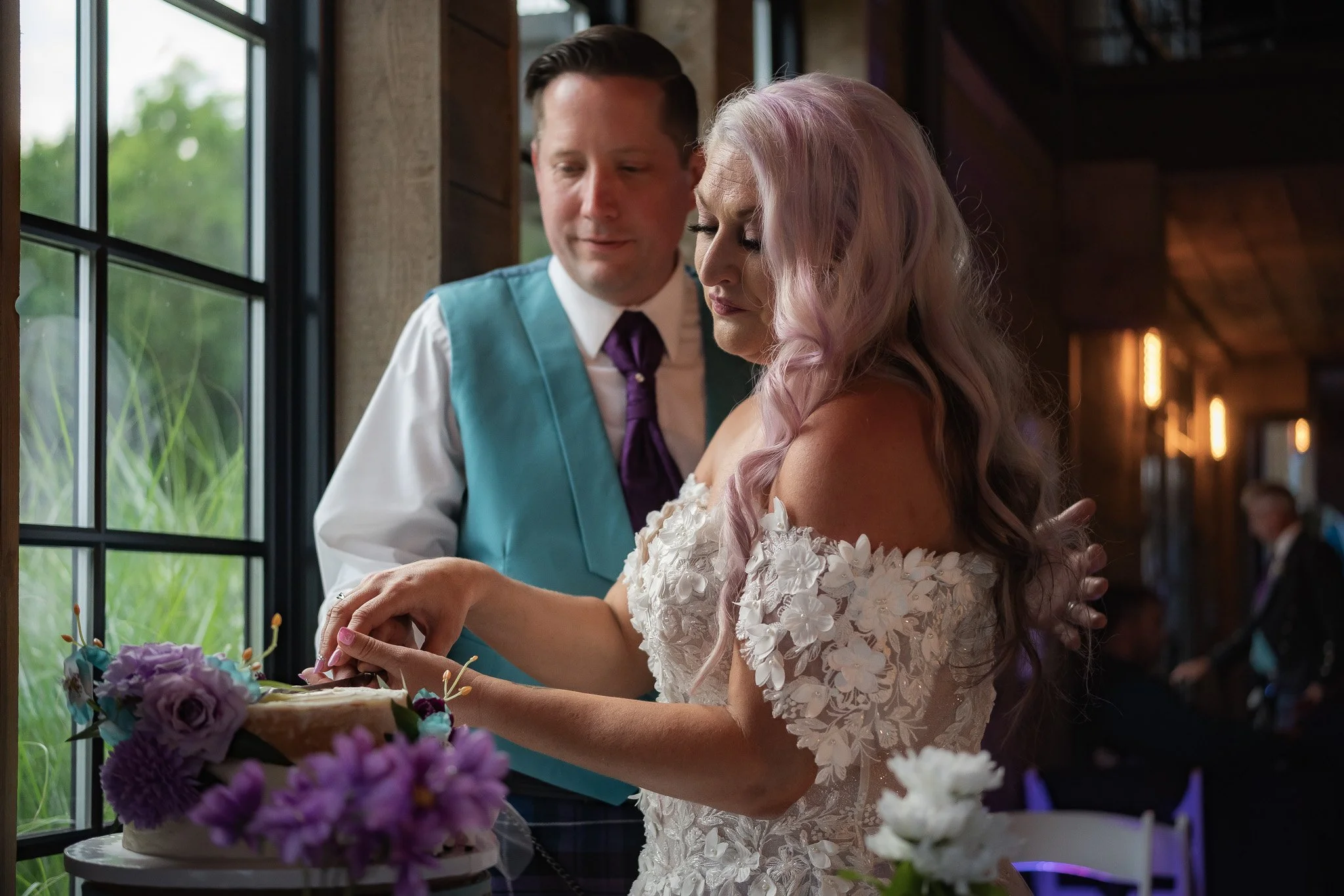 A woman with pastel pink wavy hair in an off-shoulder white floral dress is cutting a wedding cake decorated with purple and white flowers, while a man in a blue vest and purple tie looks on.