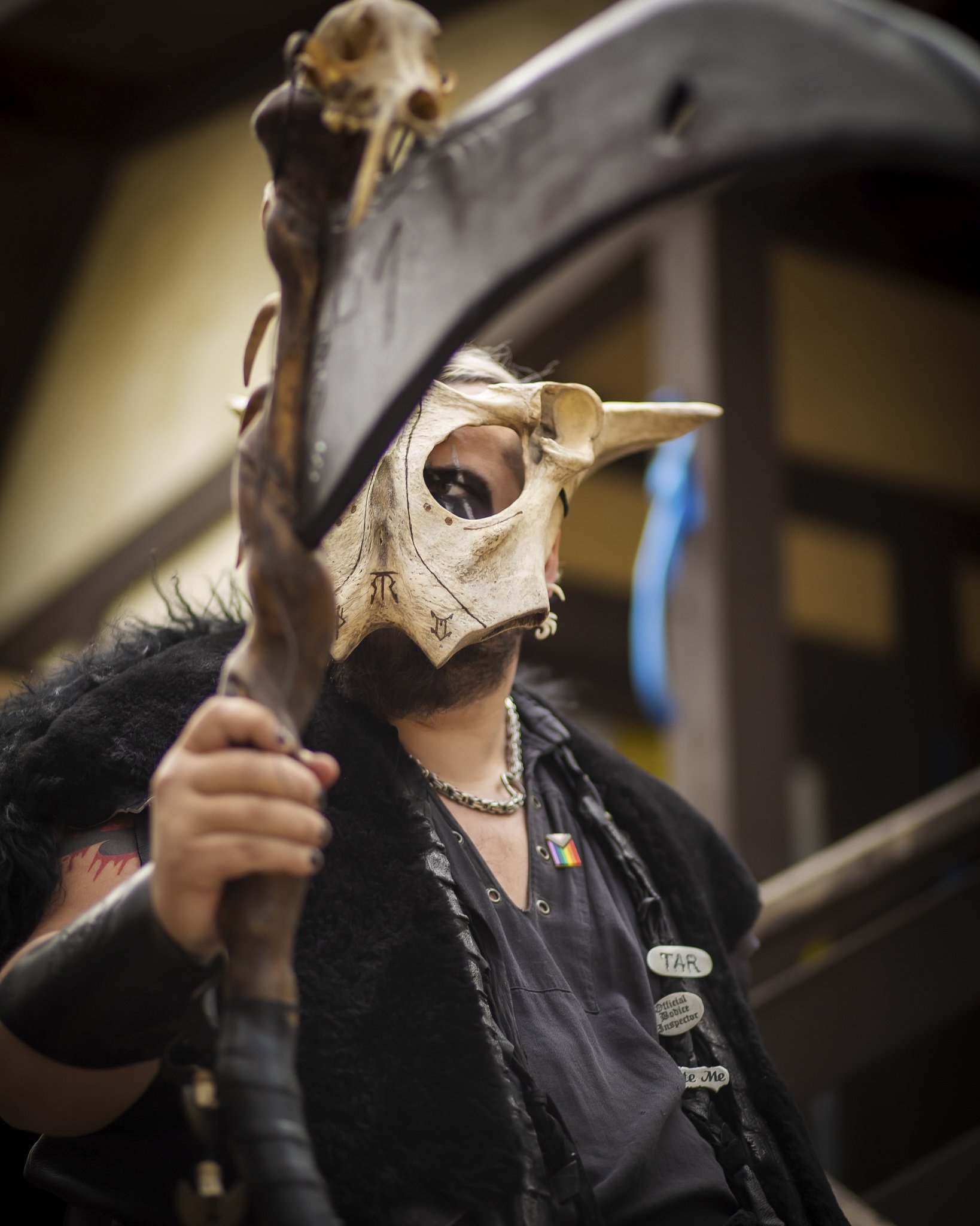Person wearing a tribal mask with a animal skull and holding a staff, dressed in black clothing with various badges and jewelry.