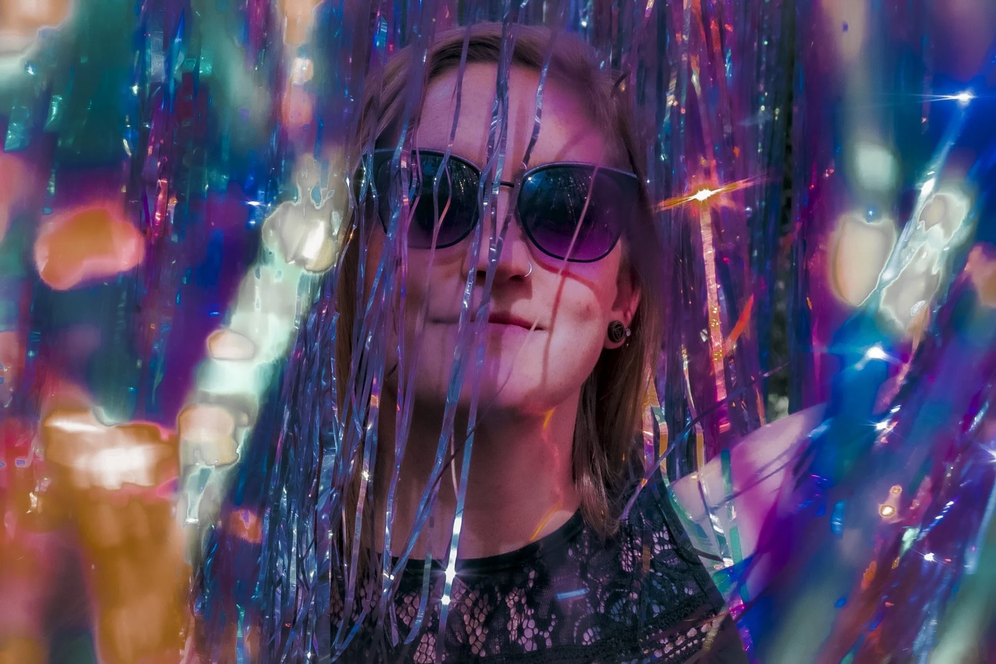 Person with short brown hair, wearing sunglasses, and a nose ring, standing behind colorful metallic streamers.