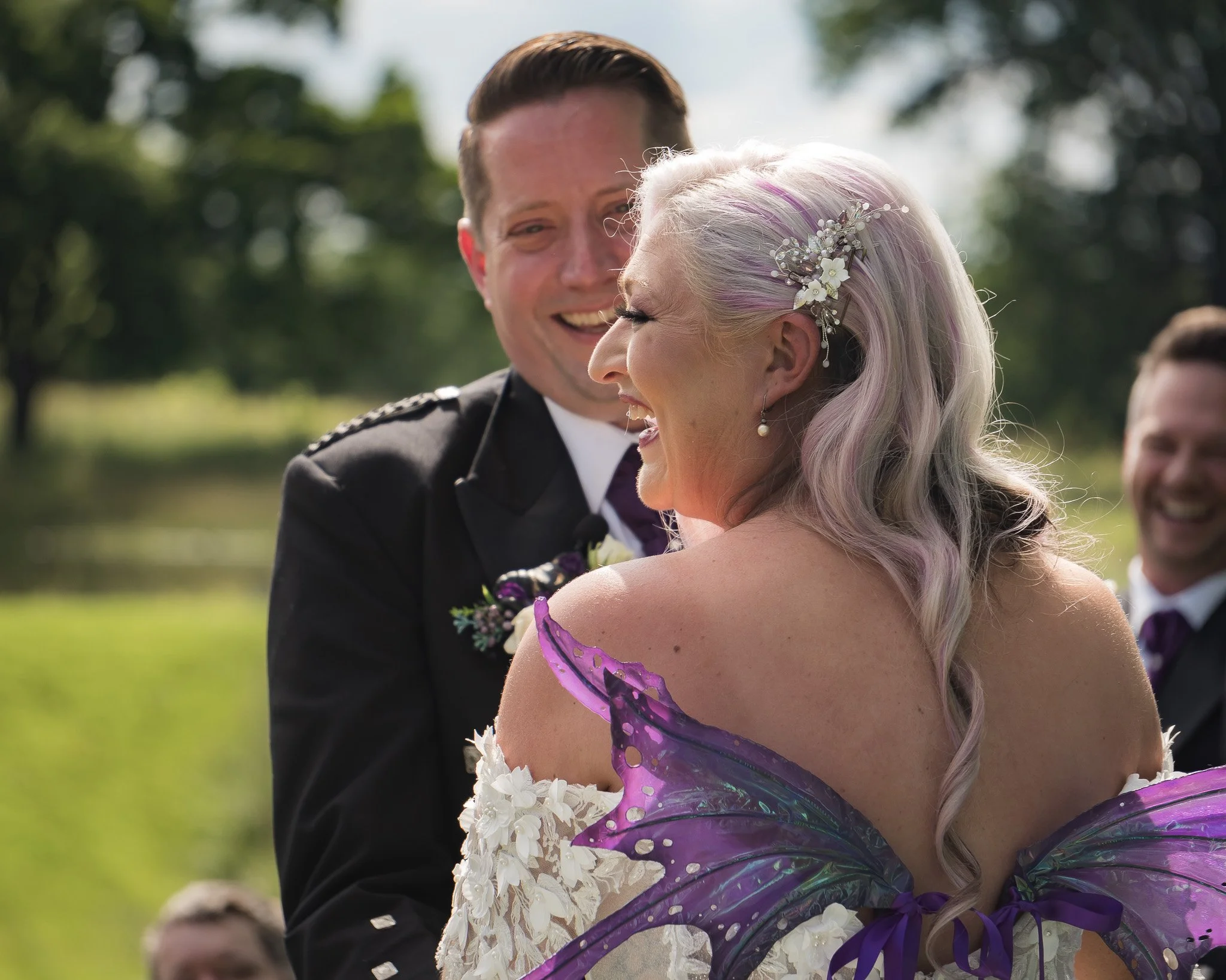 A bride with purple butterfly wings and a groom in a black tuxedo smiling at each other during a wedding outdoors, with a blurred background of trees and grass.
