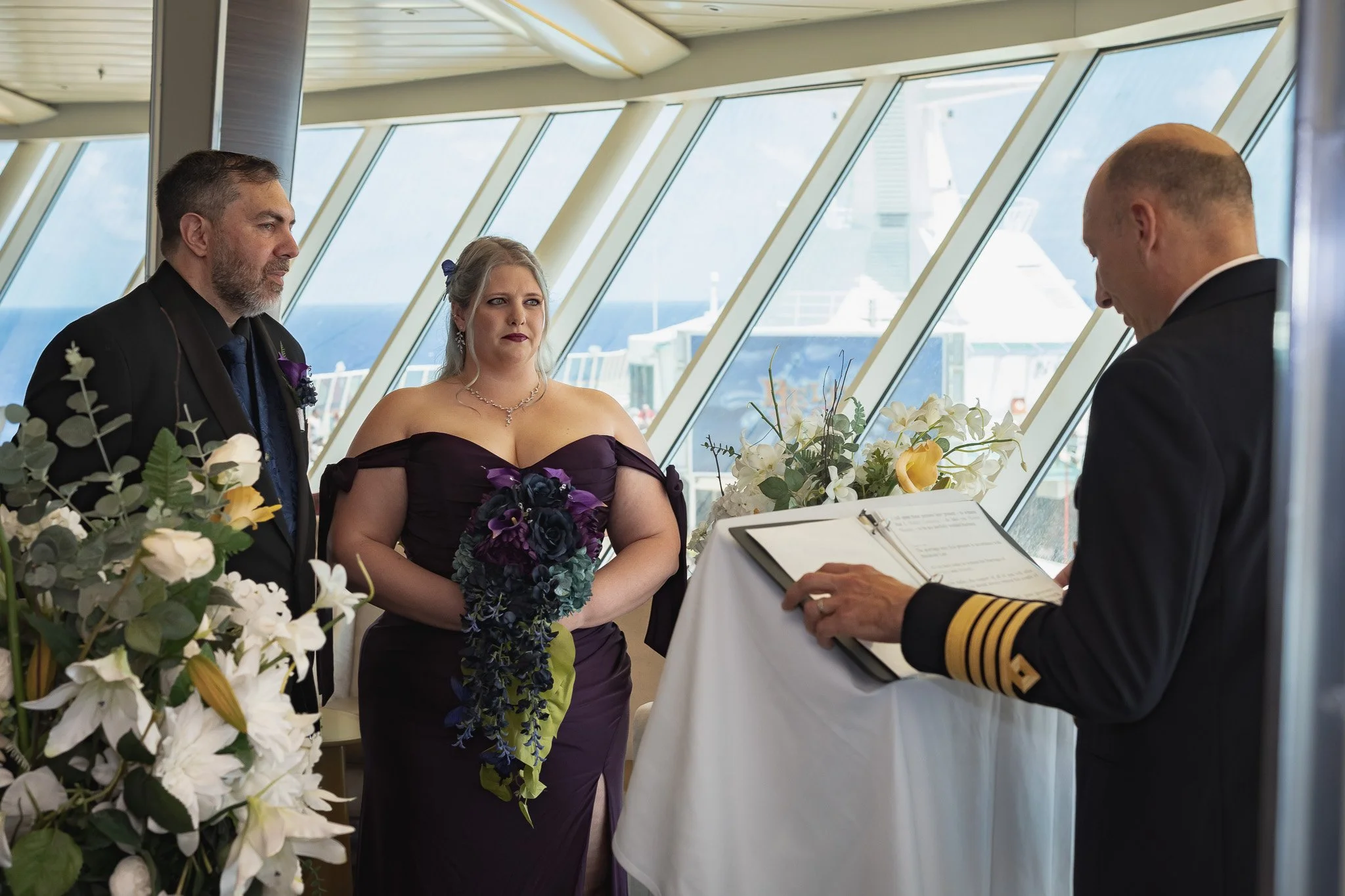 A couple stands before a captain during a wedding ceremony inside a ship's reception area, with large windows showing the sea outside. The woman wears a dark purple gown and holds a floral bouquet, while the man wears a dark suit. The captain wears a uniform and is reading from a book.
