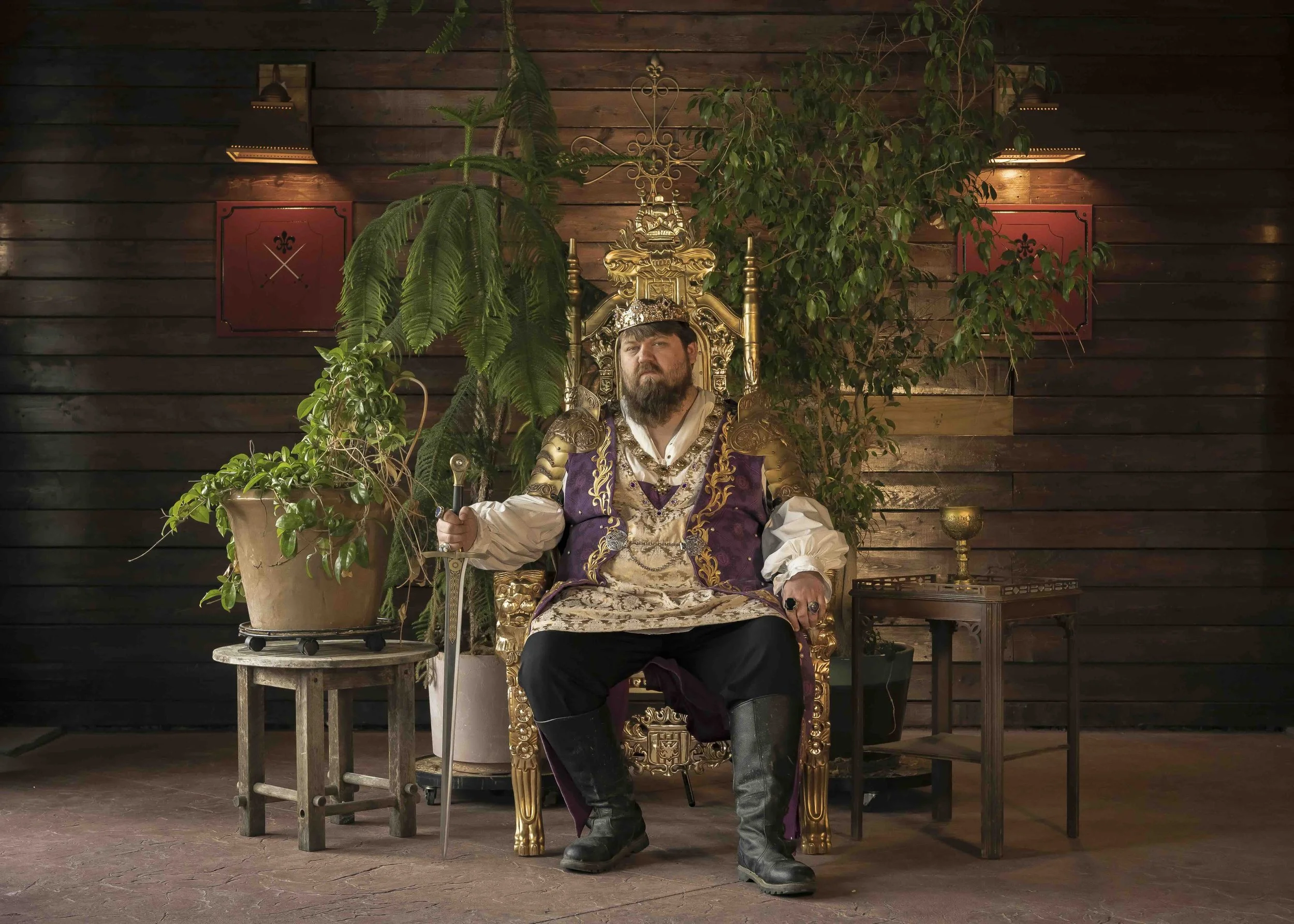 A man dressed as a king sitting on a golden throne, wearing a crown and holding a sword. The background features wooden walls, potted plants, and side tables.