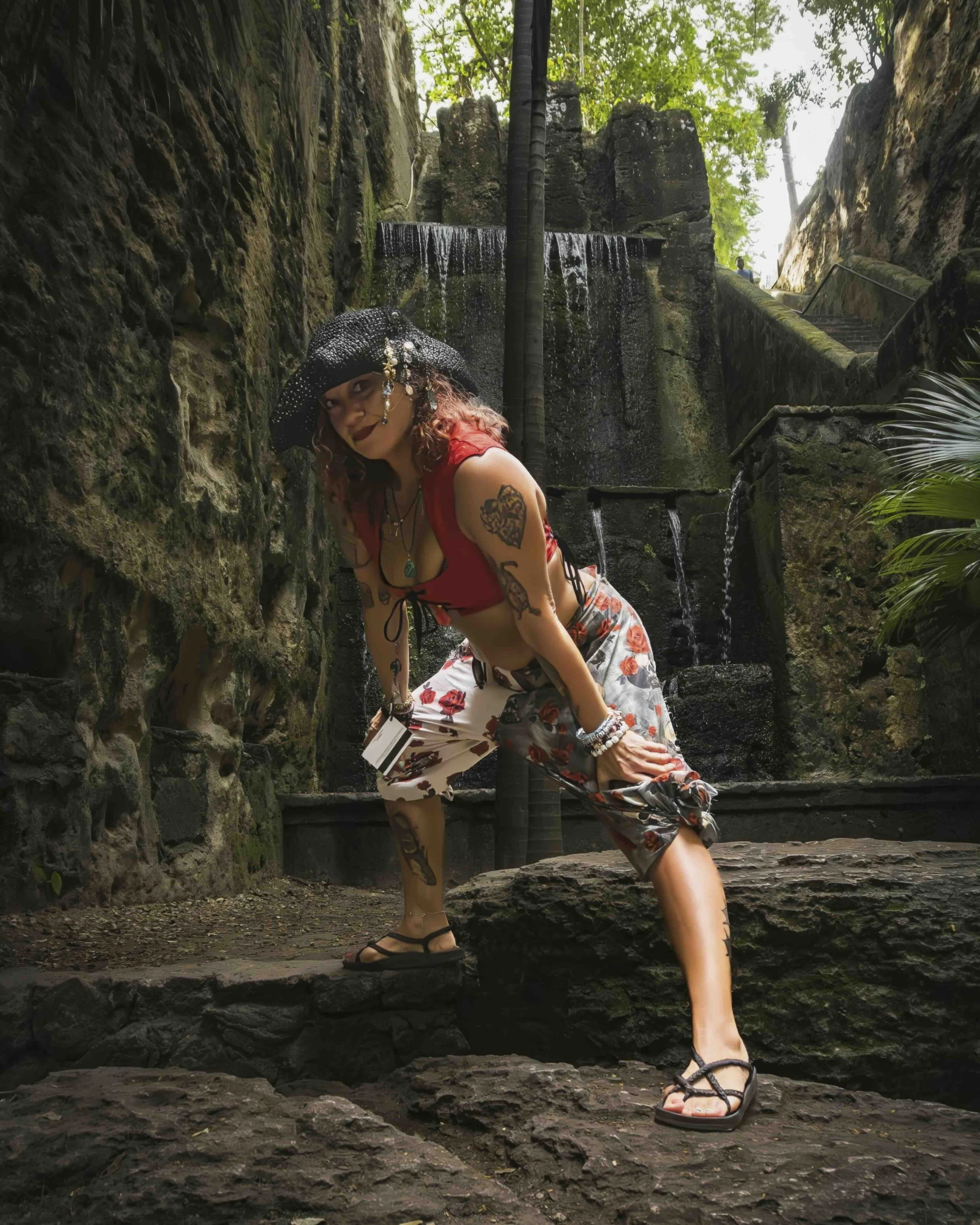 A woman with tattoos wearing a black hat with decorative beads, red sleeveless top, floral patterned pants, and black sandals, posing with one foot on a rock in front of a waterfall in a lush, green forest.