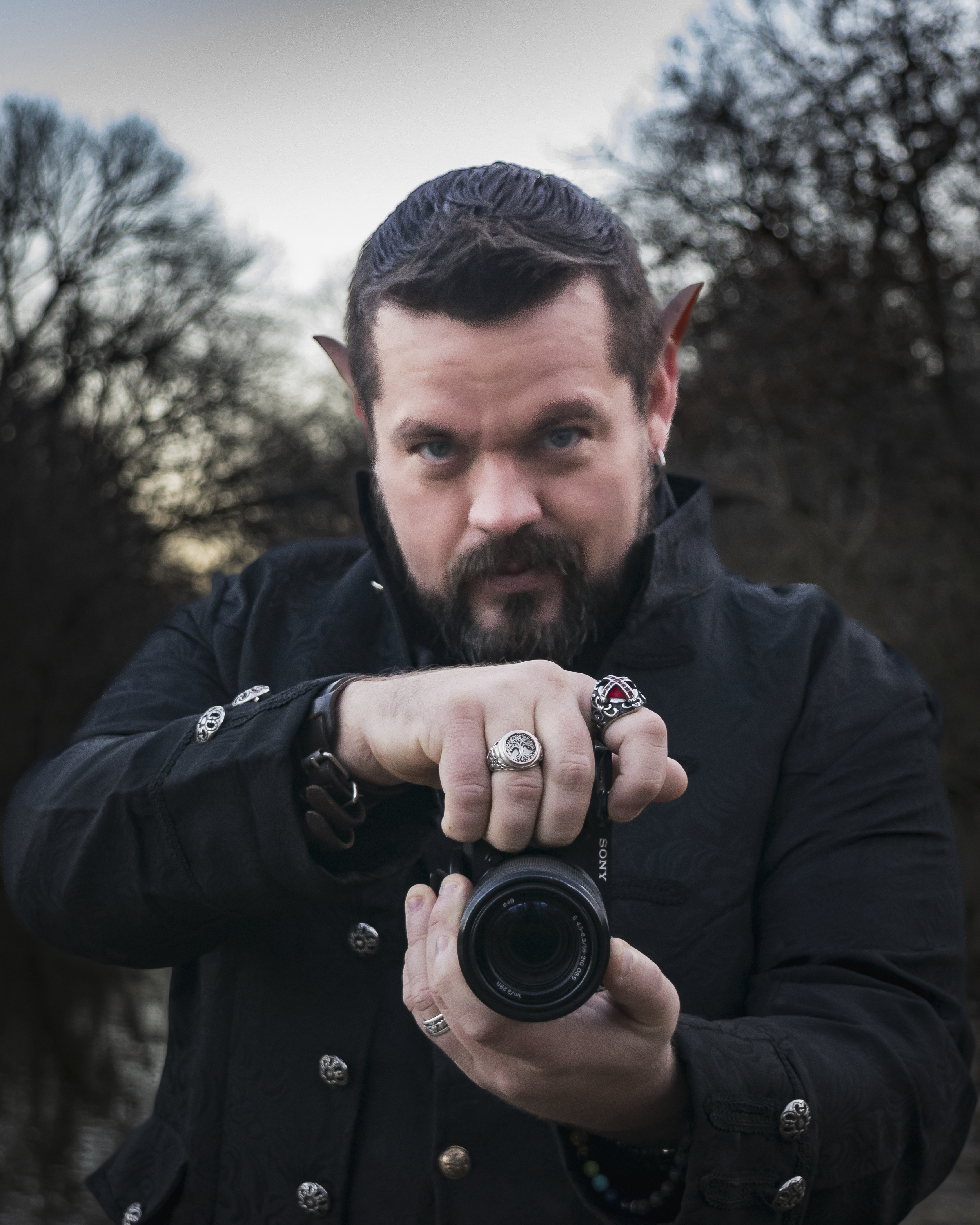 The Faetographer holding a camera, wearing rings and rings, standing outdoors during dusk with bare trees in the background.
