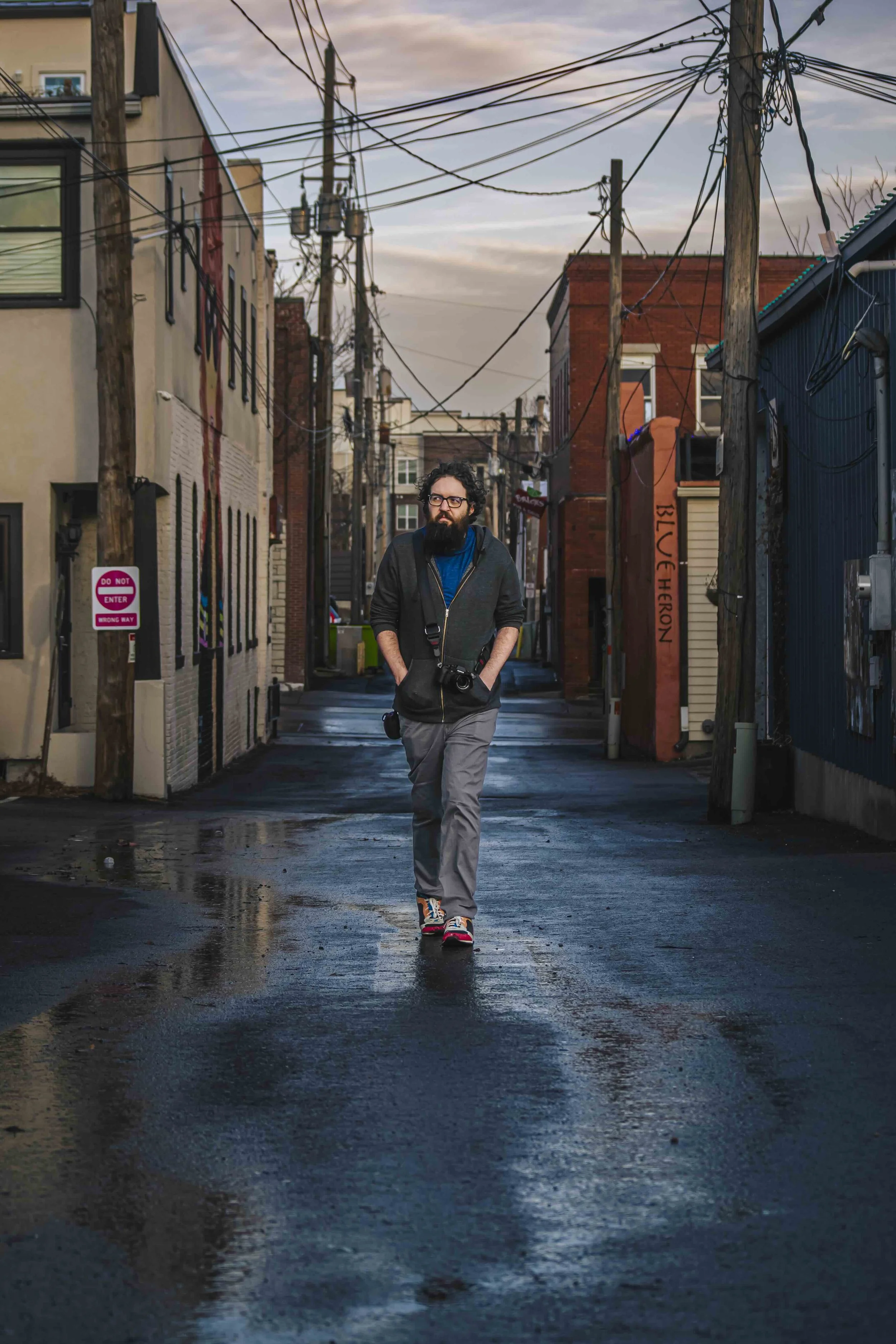 A man walking down a wet urban alleyway at sunset, carrying a camera with a strap around his neck, with buildings and power lines overhead.