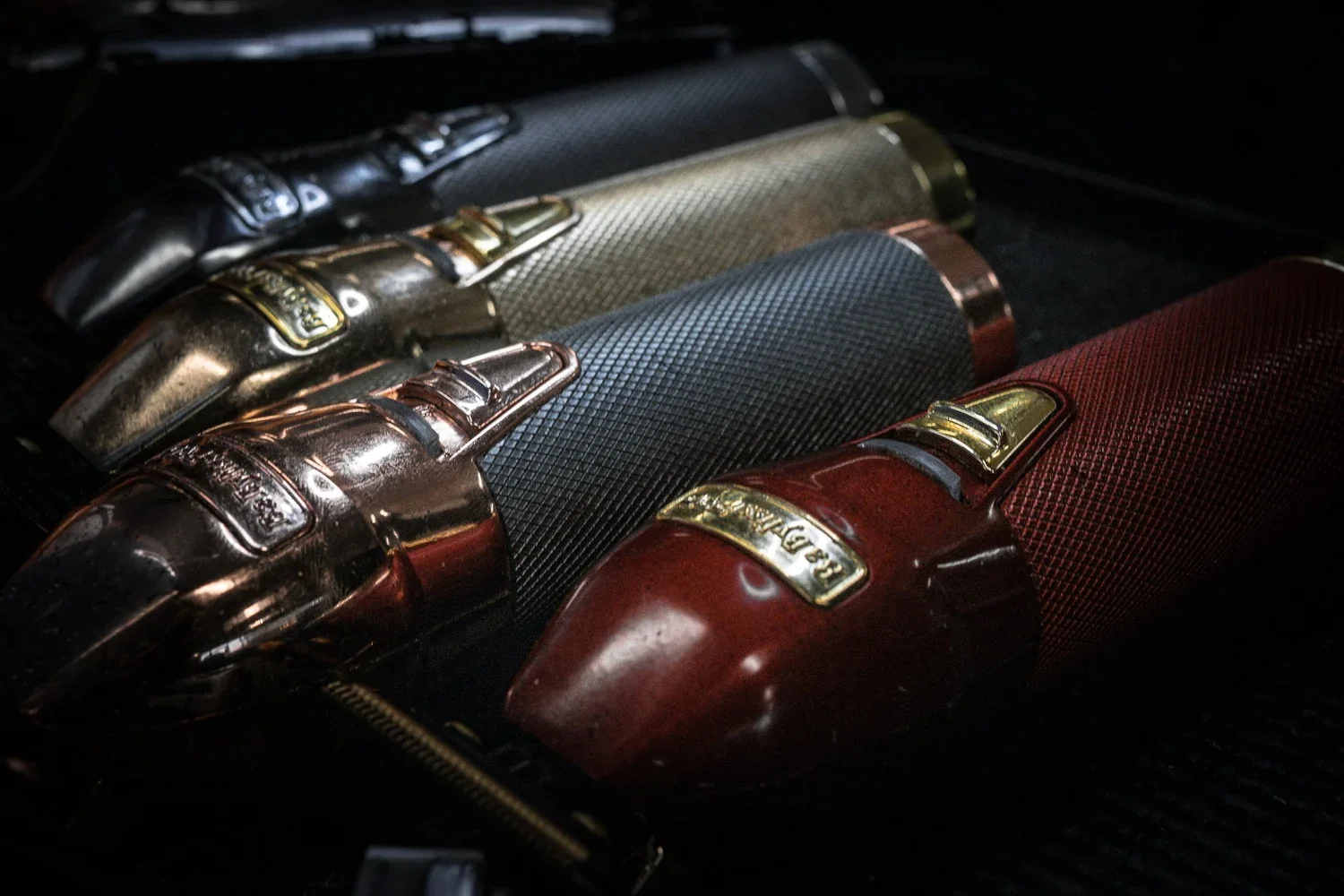 A row of luxury hair trimmer with metallic finishes and textured grips, mostly in dark tones with one in red, arranged on a black surface.