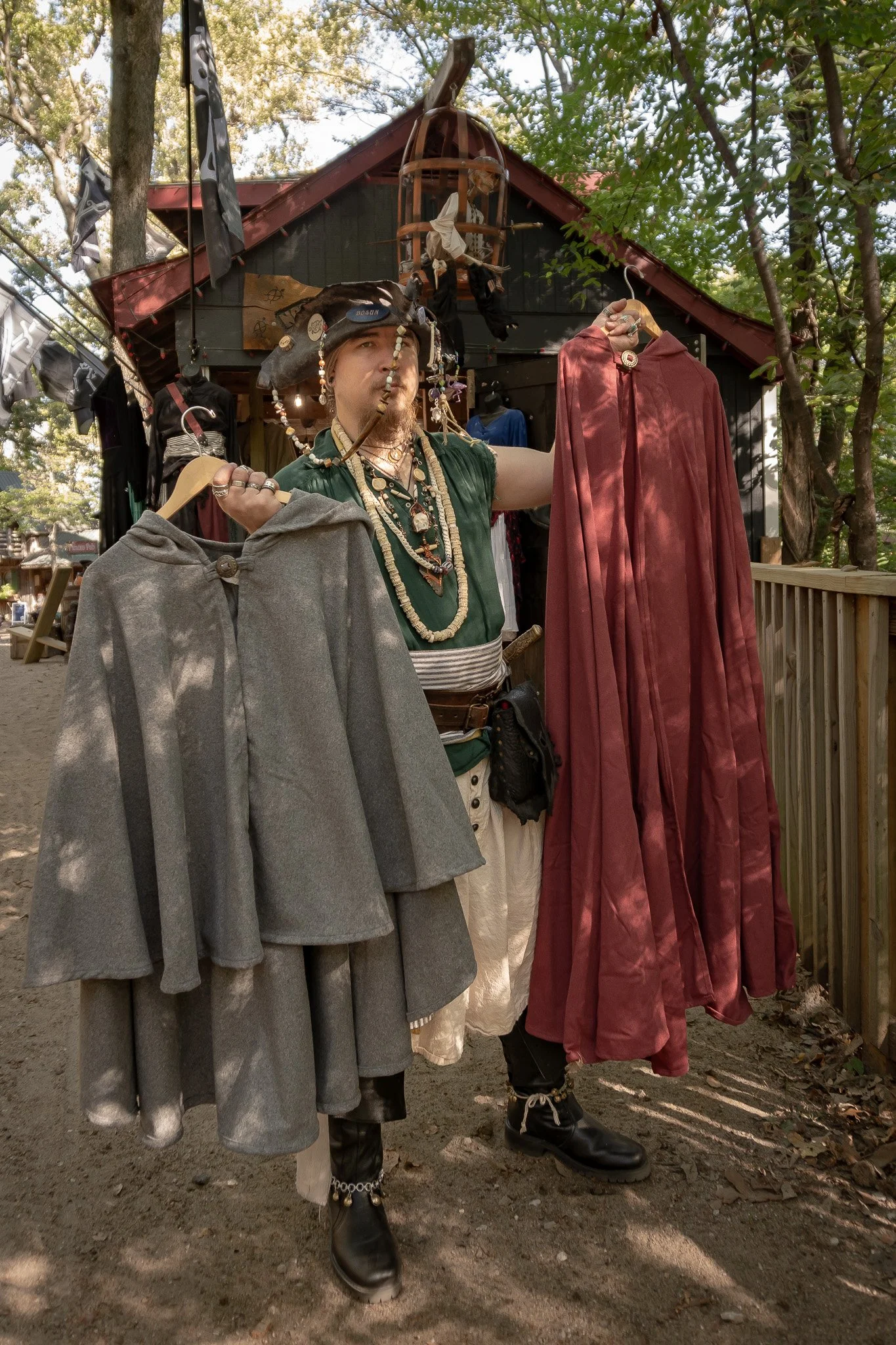 A person dressed as a pirate with facial jewelry and necklaces, standing in front of a small shop with pirate-themed accessories and clothing, holding a gray and a red cape.