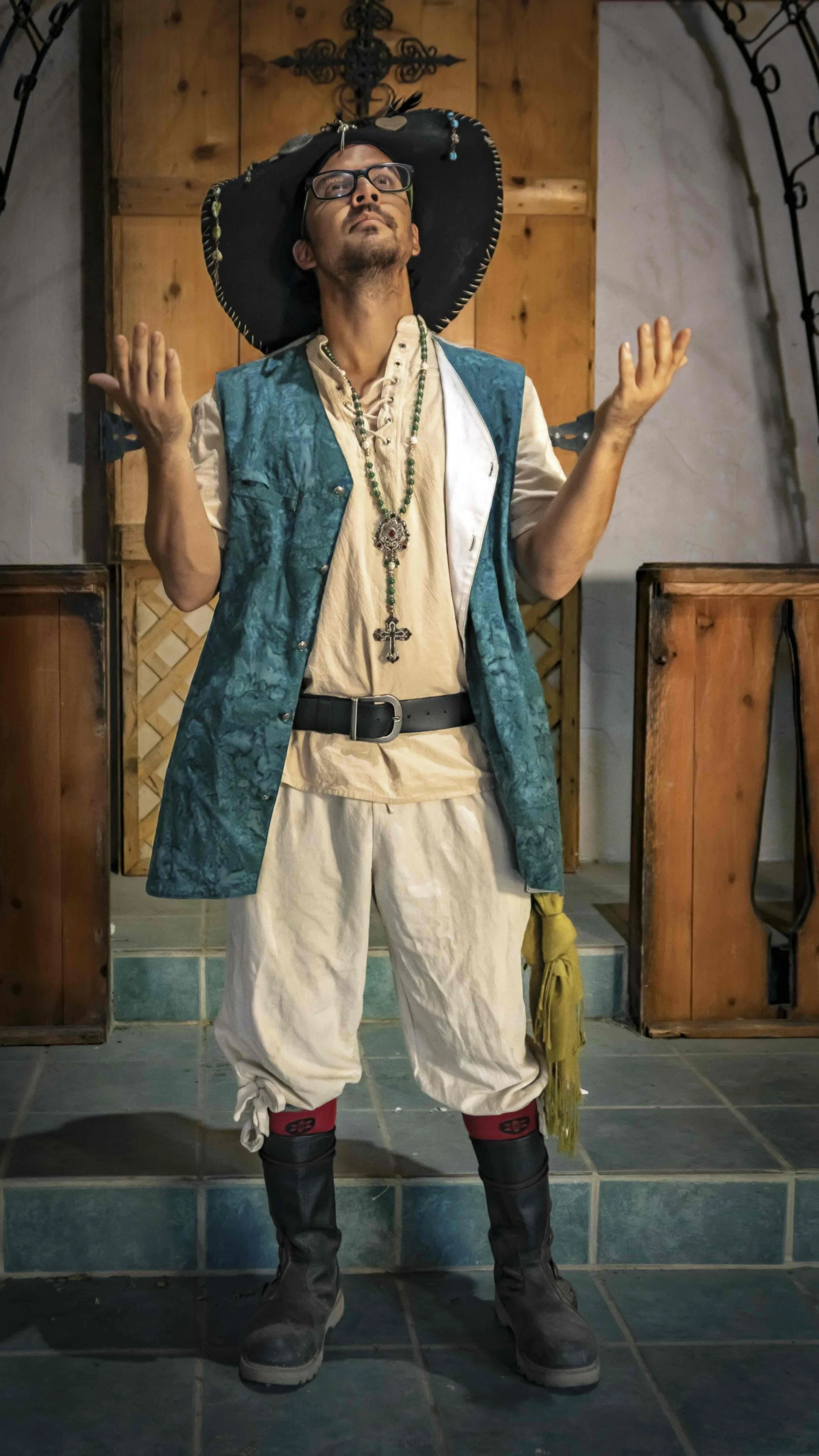 A man dressed in a colorful, theatrical costume standing with arms raised, wearing a large black hat with decorations, glasses, and jewelry, in front of a wooden background with a cross and tiled floor.